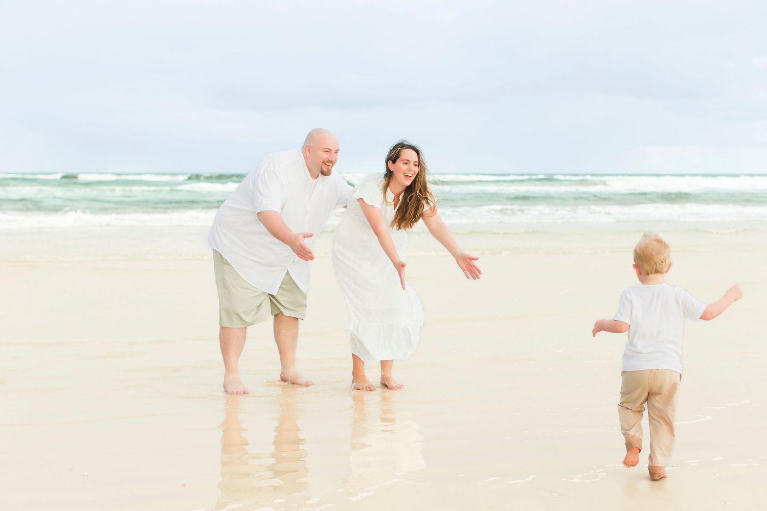 Playful family beach moment as toddler runs toward parents on a bright sandy shoreline.