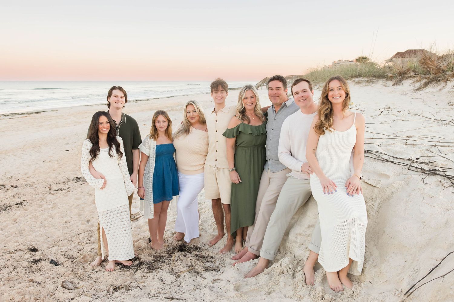 Large extended family of nine smiling together on a sandy beach at pastel sunset.