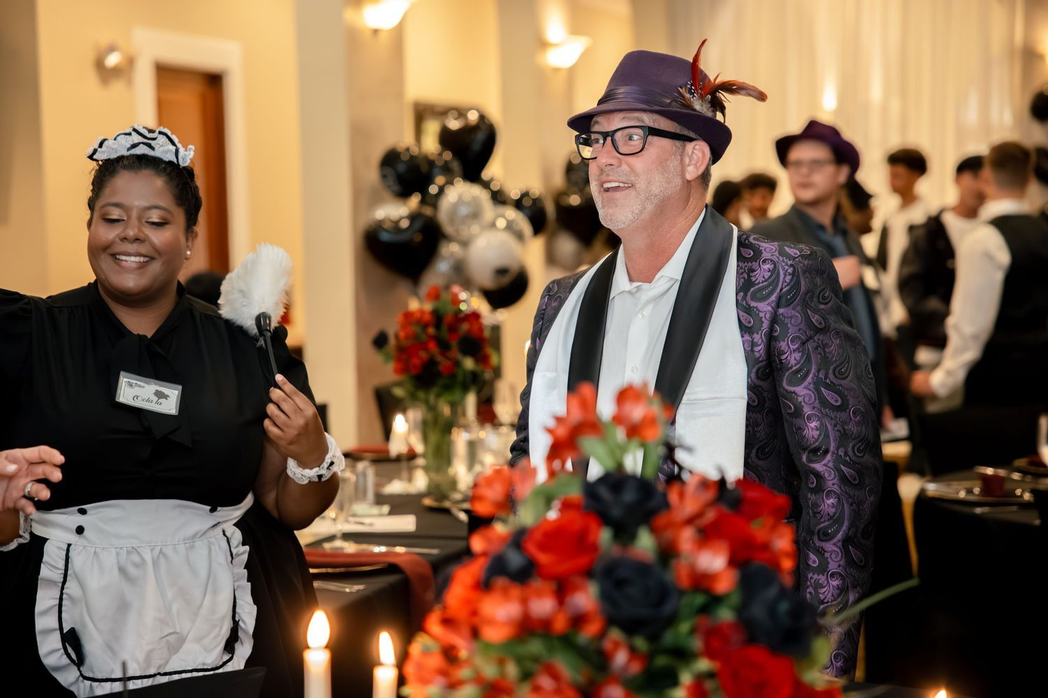 Party guests in costumes laugh near candlelit tables with red floral arrangements and black balloons at birthday event.