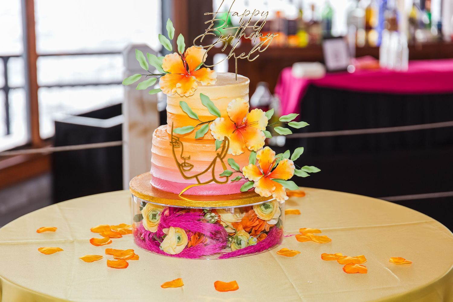 A elegant three-tier birthday cake decorated with orange hibiscus flowers on a yellow tablecloth.