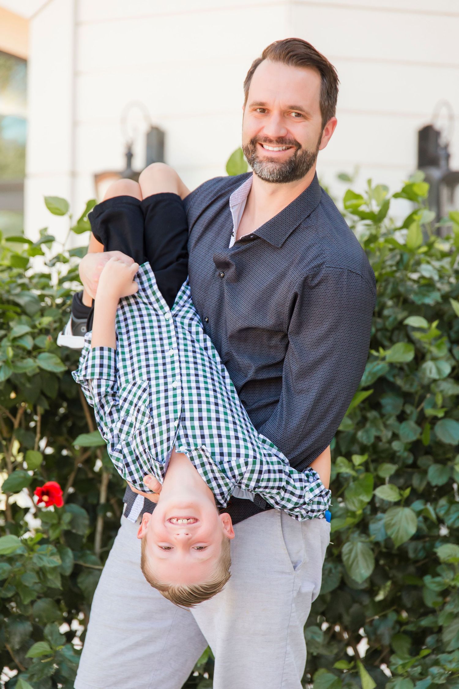 Smiling father playfully holding young son upside down in front of a lush green garden hedge.