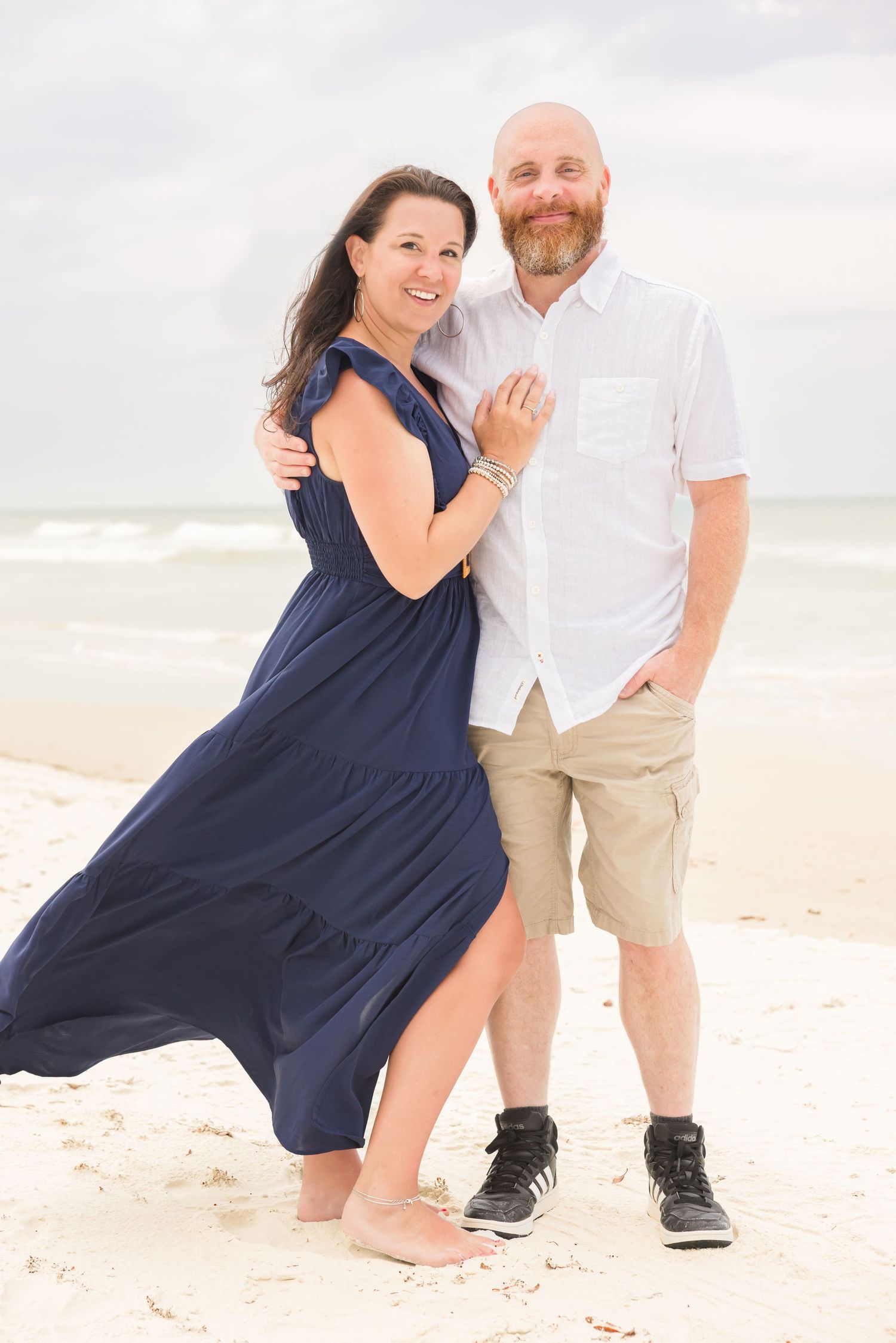 Couple embracing on a sandy beach, woman in navy dress and man in white shirt and khaki shorts.