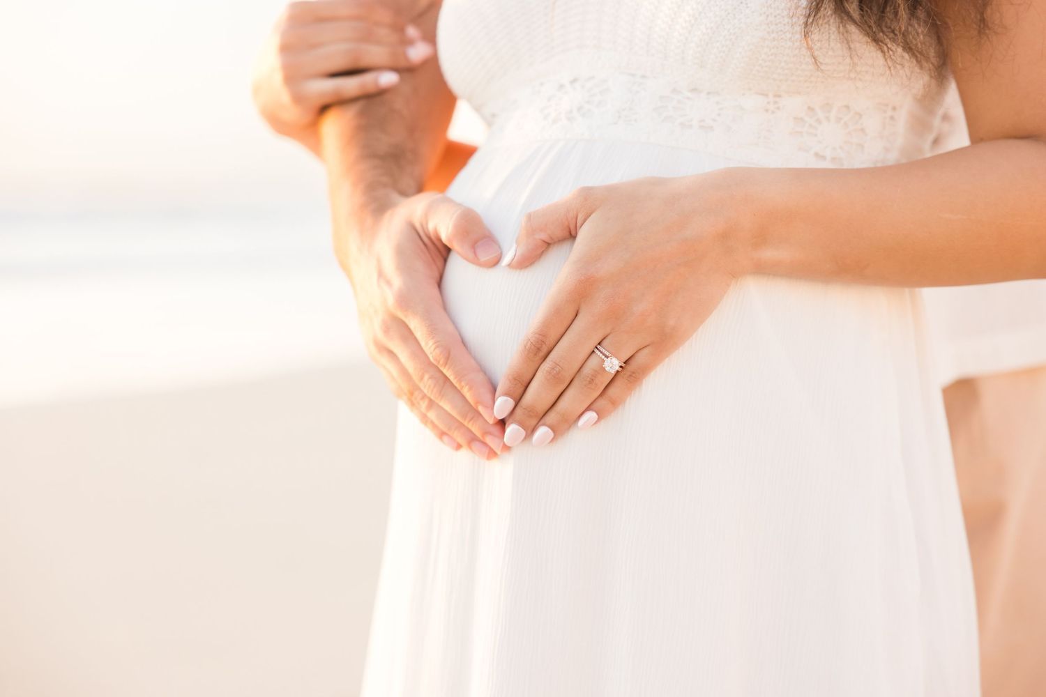 Couple forming heart shape with hands on pregnant belly, wearing wedding rings, in white dress at beach.