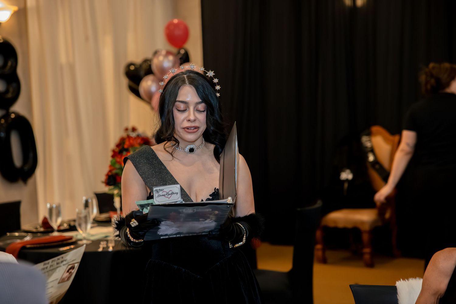 Woman in black dress and gloves reads at table during elegantly decorated 30th birthday party event.