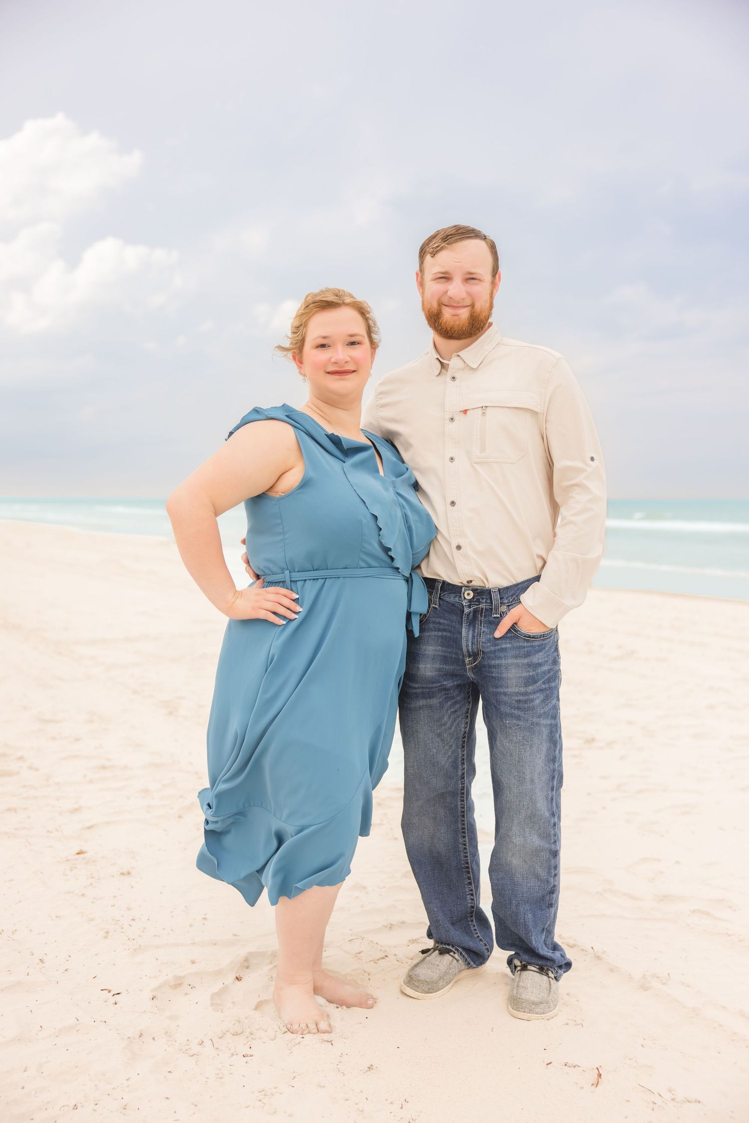 A couple stands together on a white sandy beach, woman in blue dress and man in jeans.