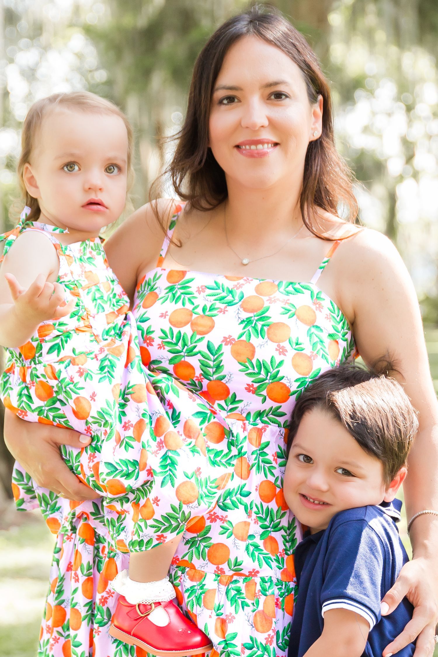 Mother and two young children wearing matching orange floral print dresses outdoors in a park.