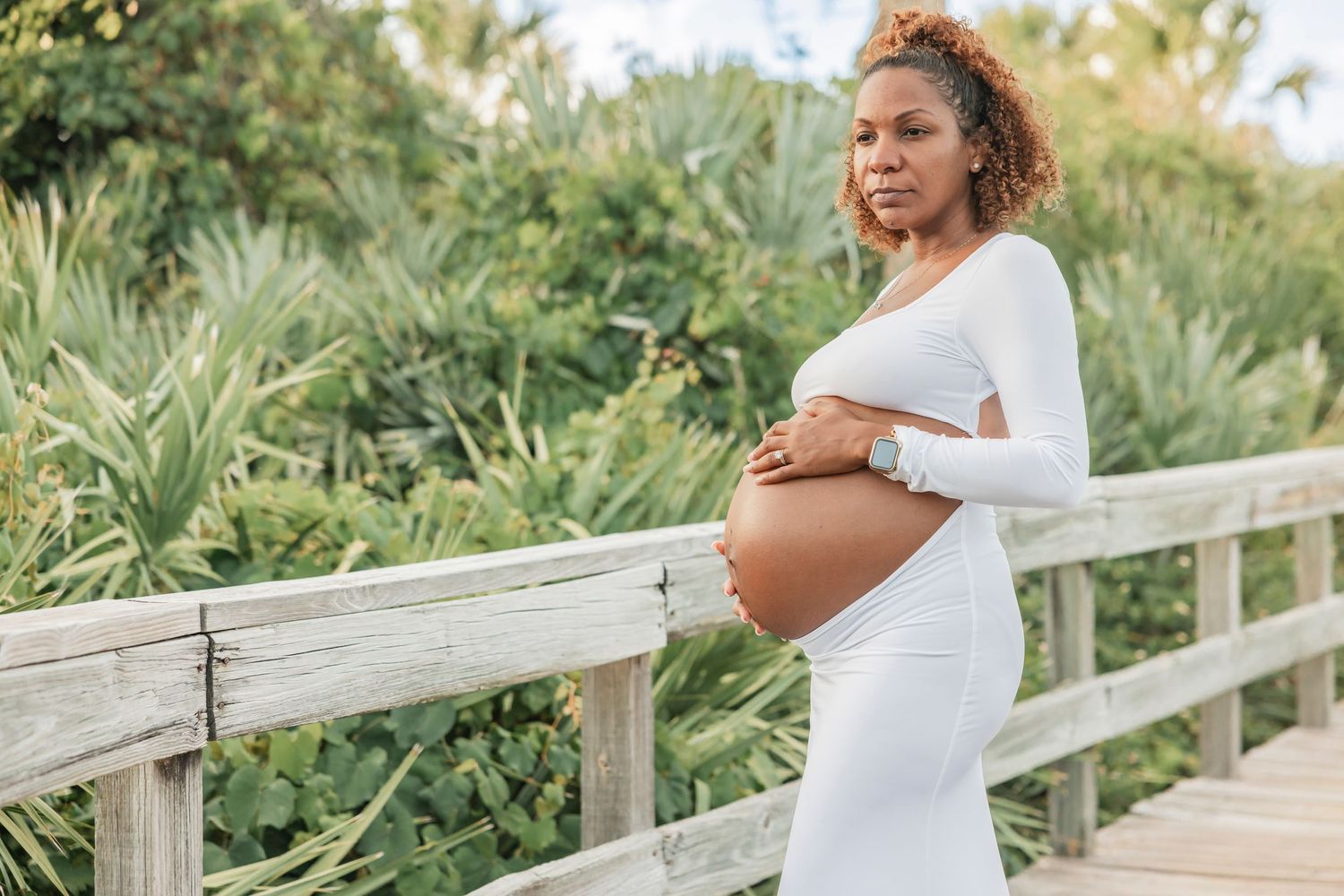 Pregnant woman in white dress standing on a wooden boardwalk, gently holding her baby bump with greenery behind.