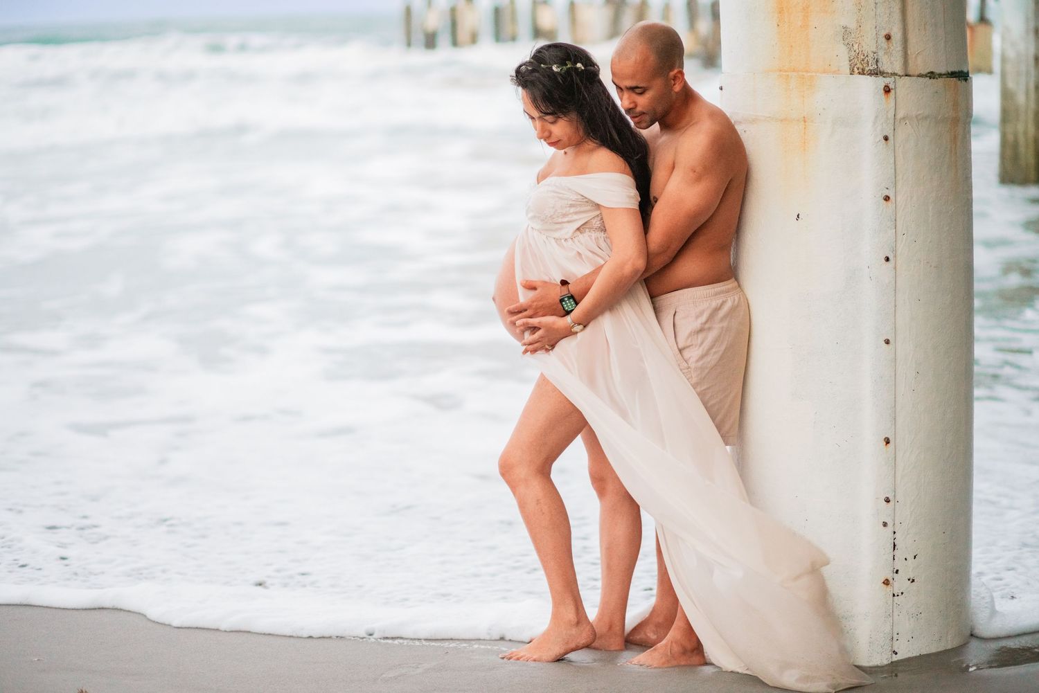 Pregnant couple leaning against pier column, white fabric flowing in breeze, beach maternity photo.