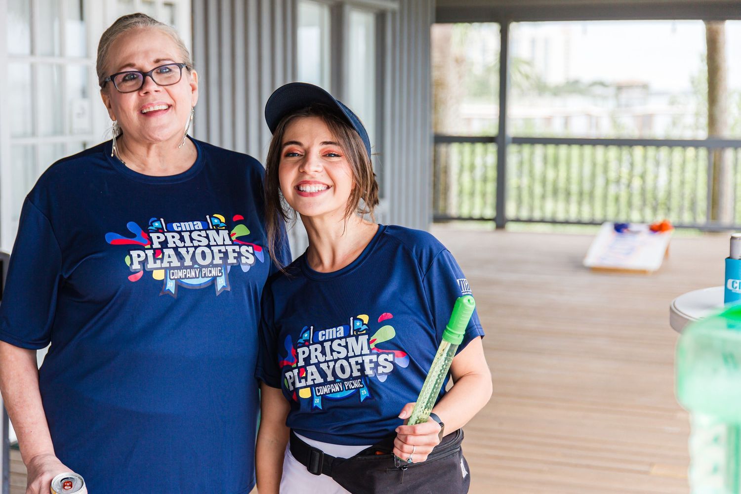 Two women in Prism Playoffs t-shirts smile together while holding green party supplies at an event.