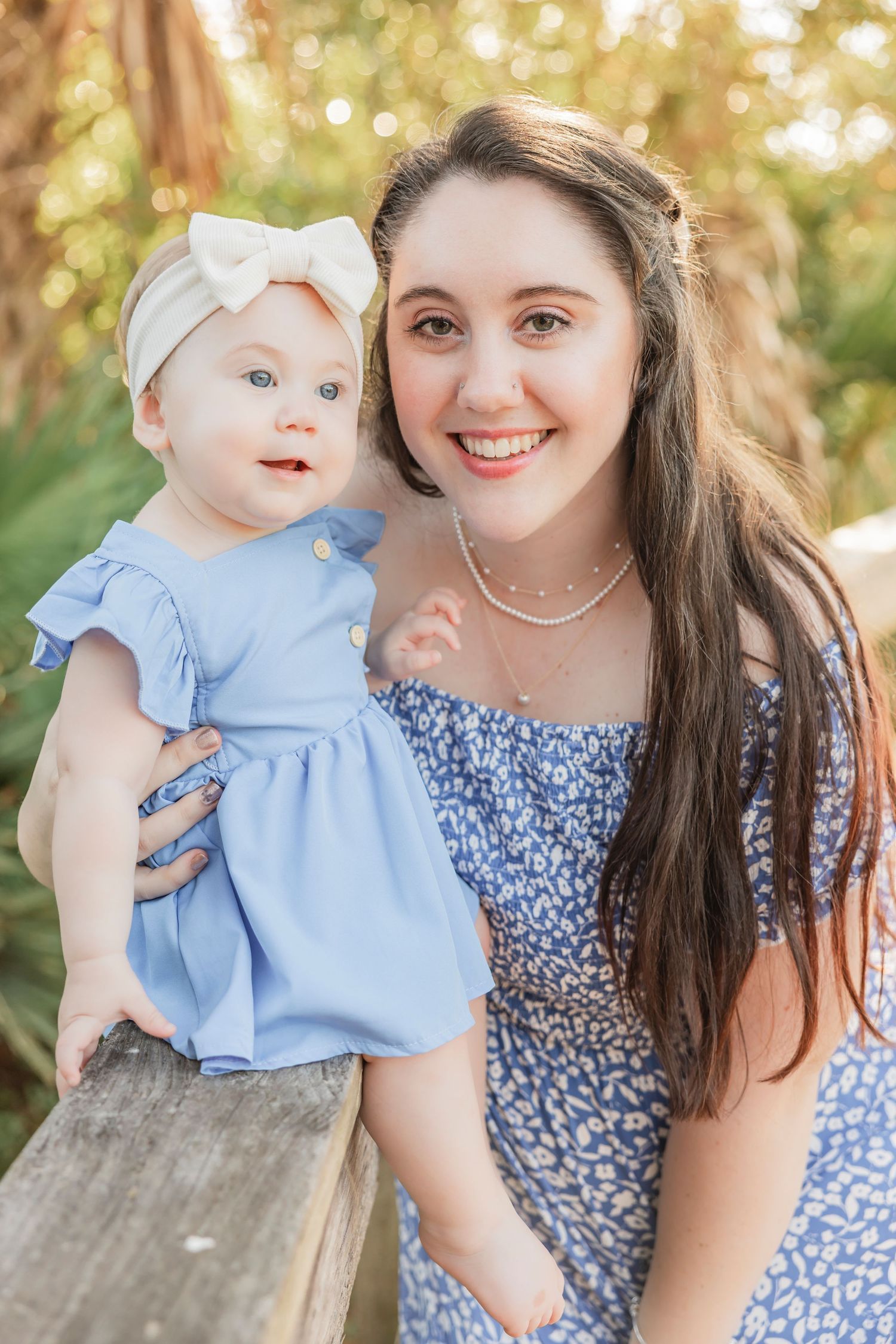 Smiling young woman holding baby girl in blue dress outdoors.