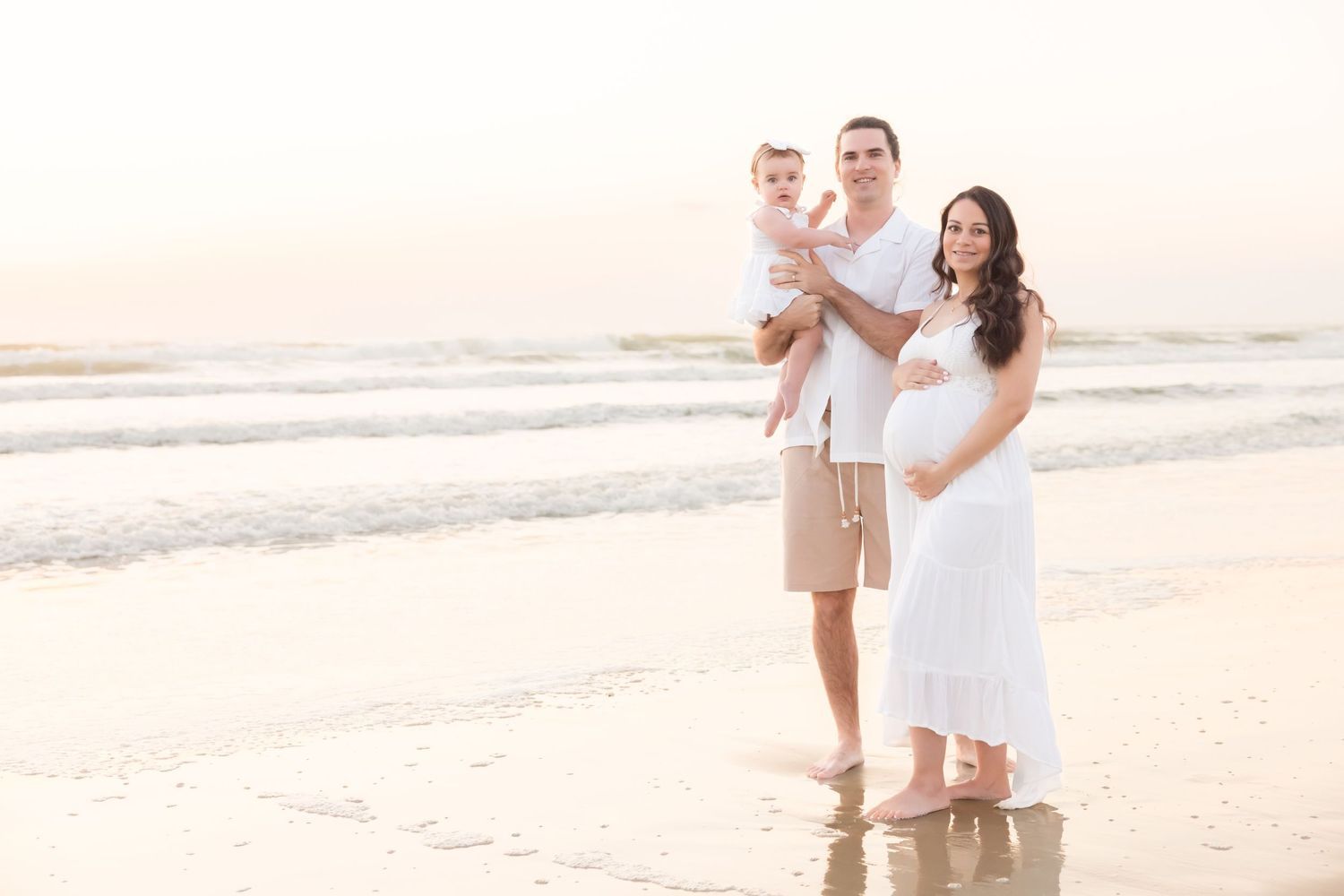 Happy family with baby and pregnant mother posing together on a sunlit sandy beach.