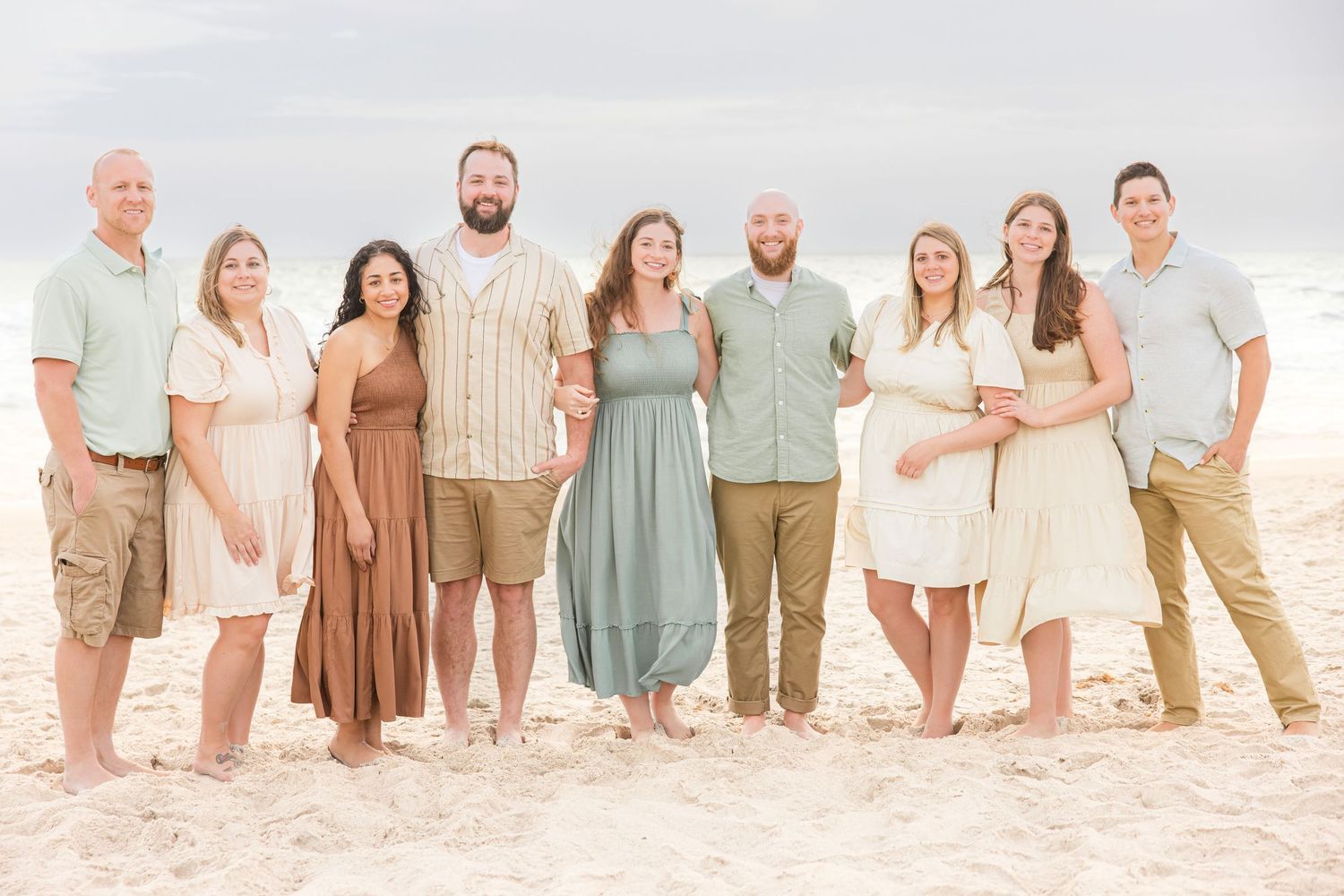 Large family group posing together on a sandy beach in coordinated neutral and pastel outfits.