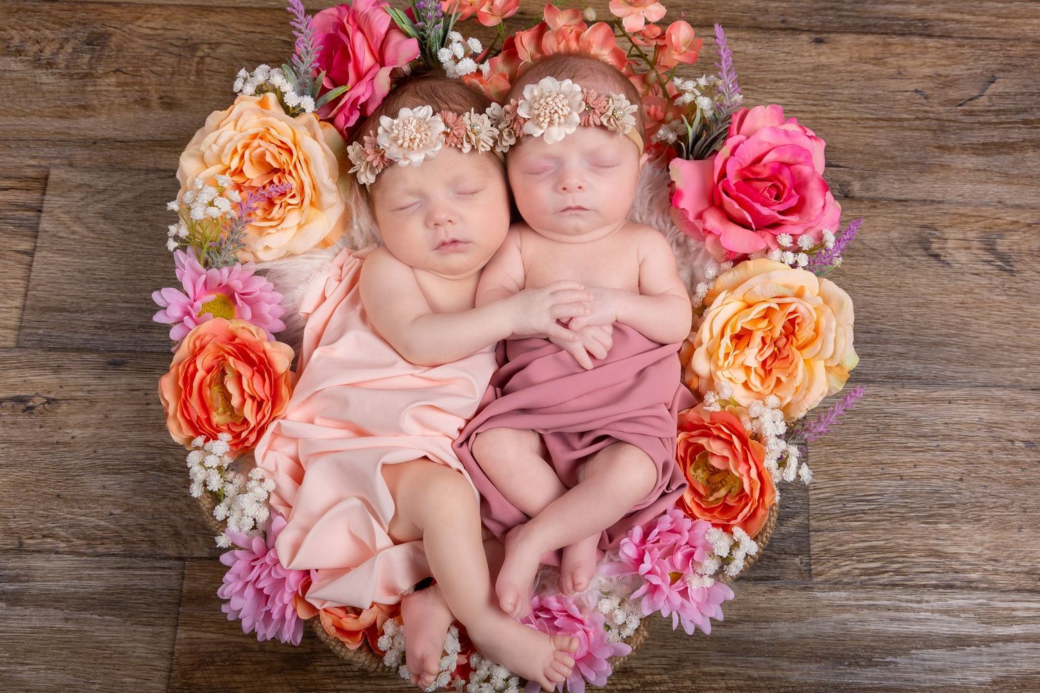Two newborn twin girls sleeping peacefully in a floral wreath arrangement with pink and orange roses.