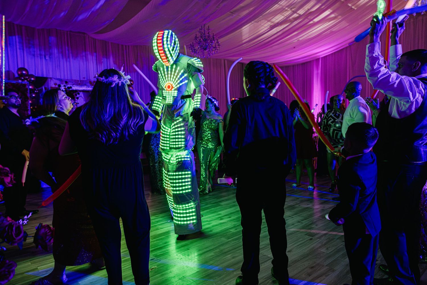 A glowing LED robot costume performer entertains guests on a dance floor at a vibrant neon-lit party event.