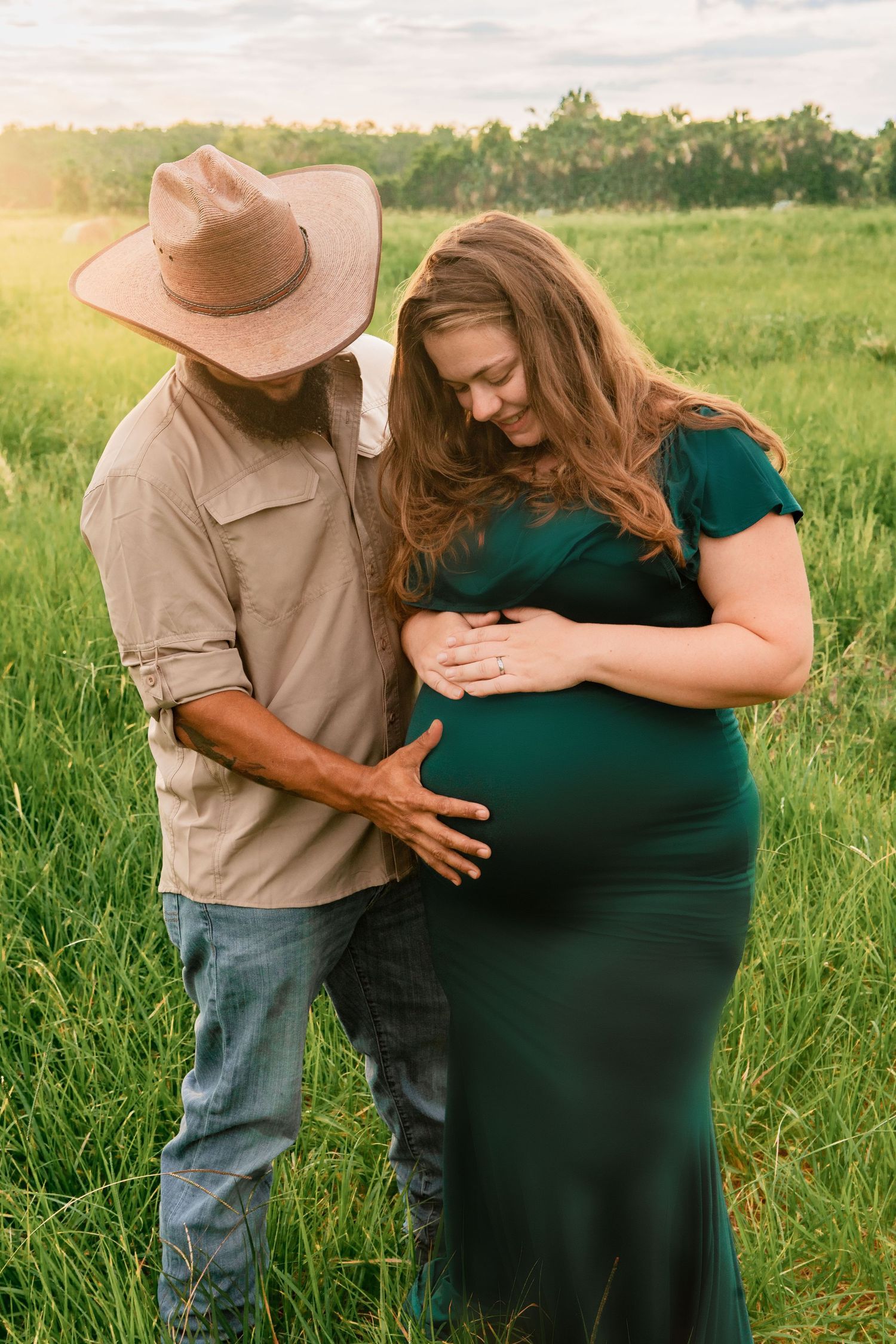 Couple in green field at golden hour, man in cowboy hat touching pregnant woman's belly.