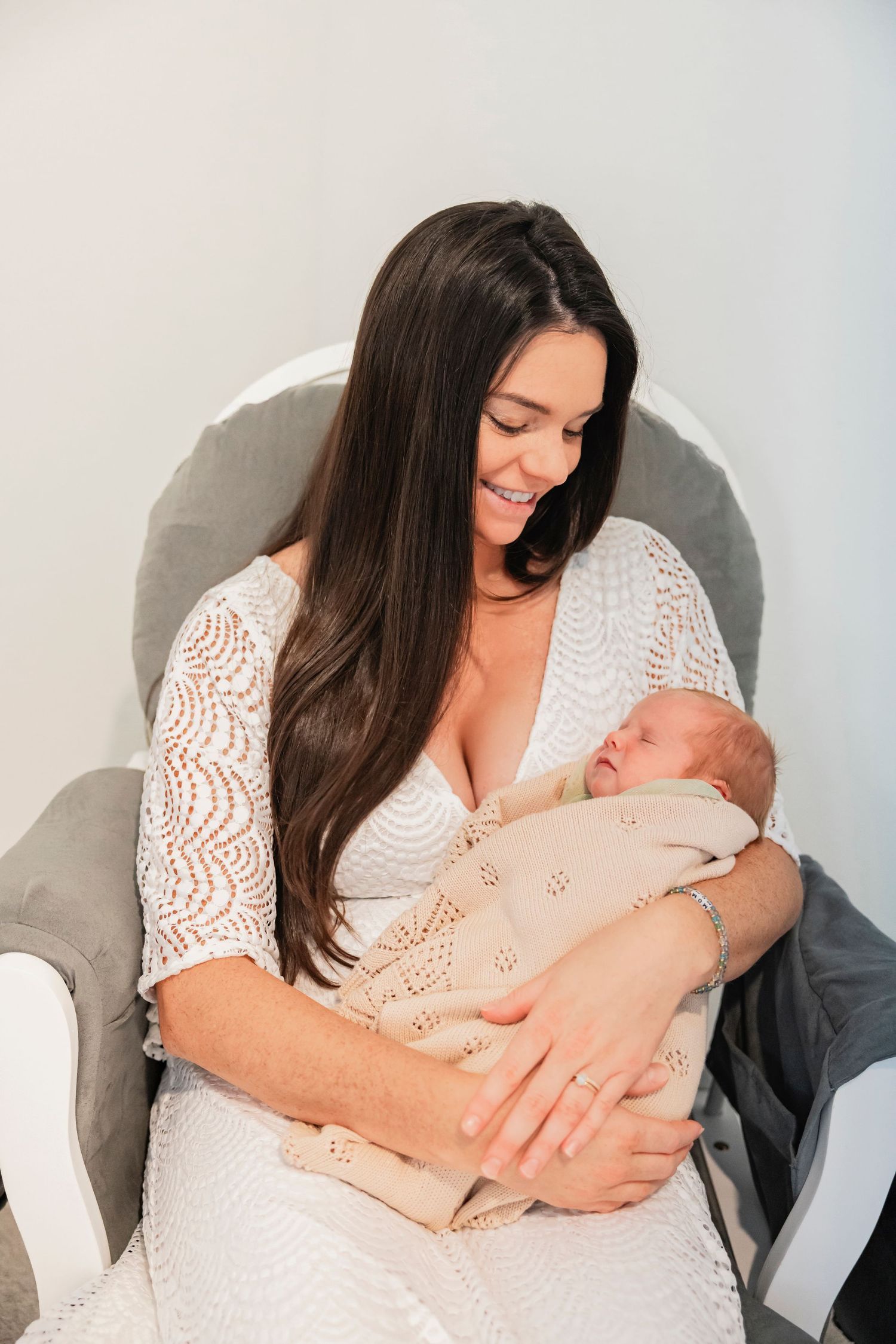 Smiling brunette mother in white lace dress cradling sleeping newborn baby wrapped in beige blanket while seated in gray chair.