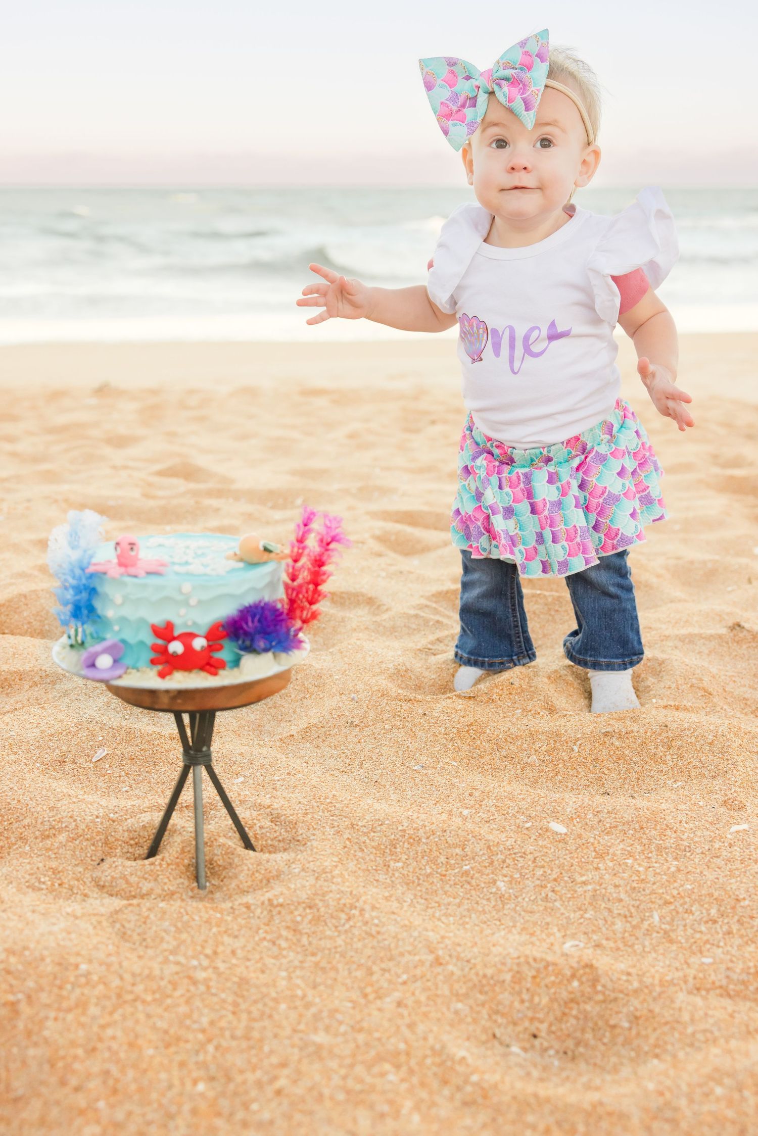 Baby girl in mermaid outfit standing near first birthday cake on beach.