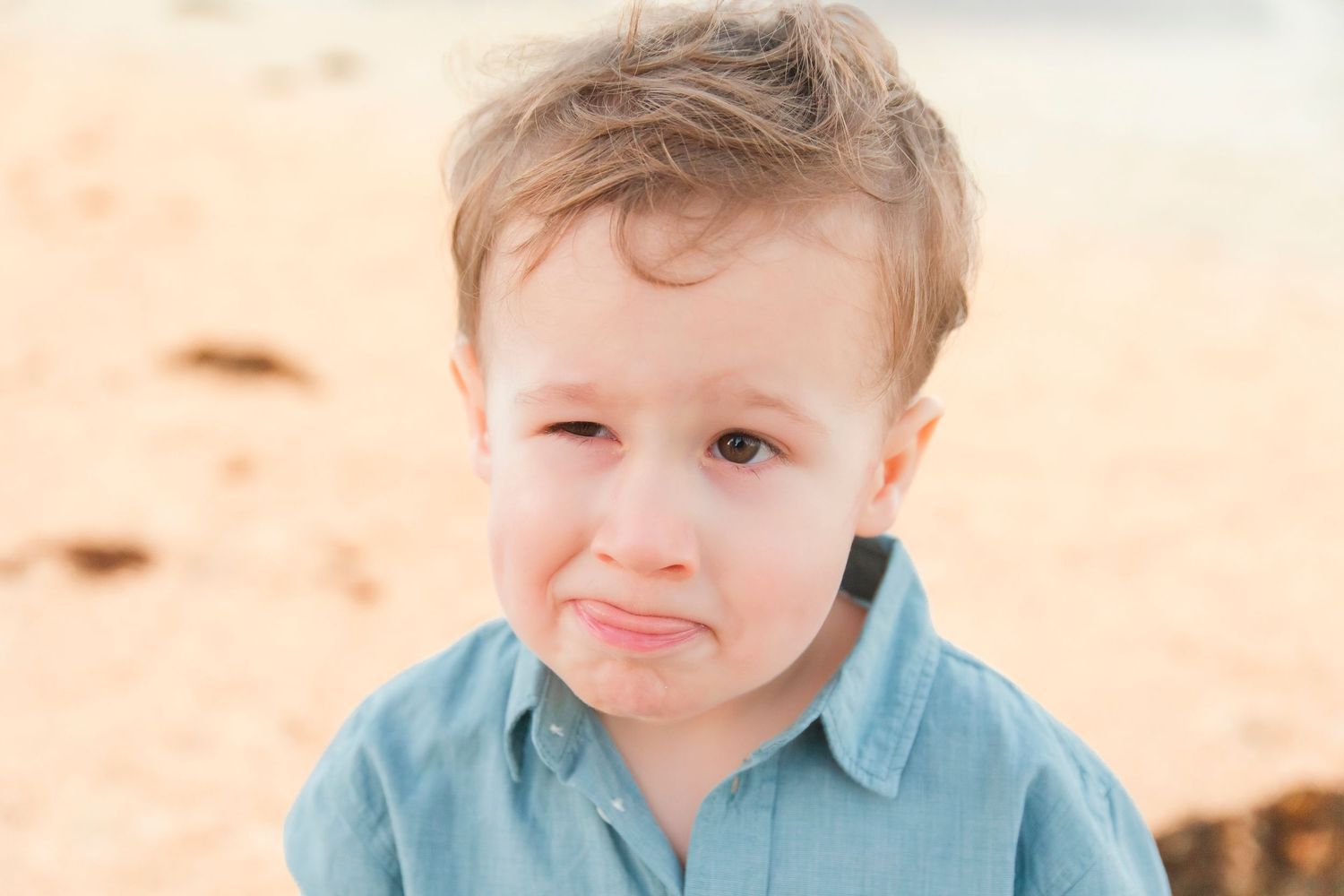 Close-up portrait of a cute toddler boy in a blue shirt making a funny face on the beach.