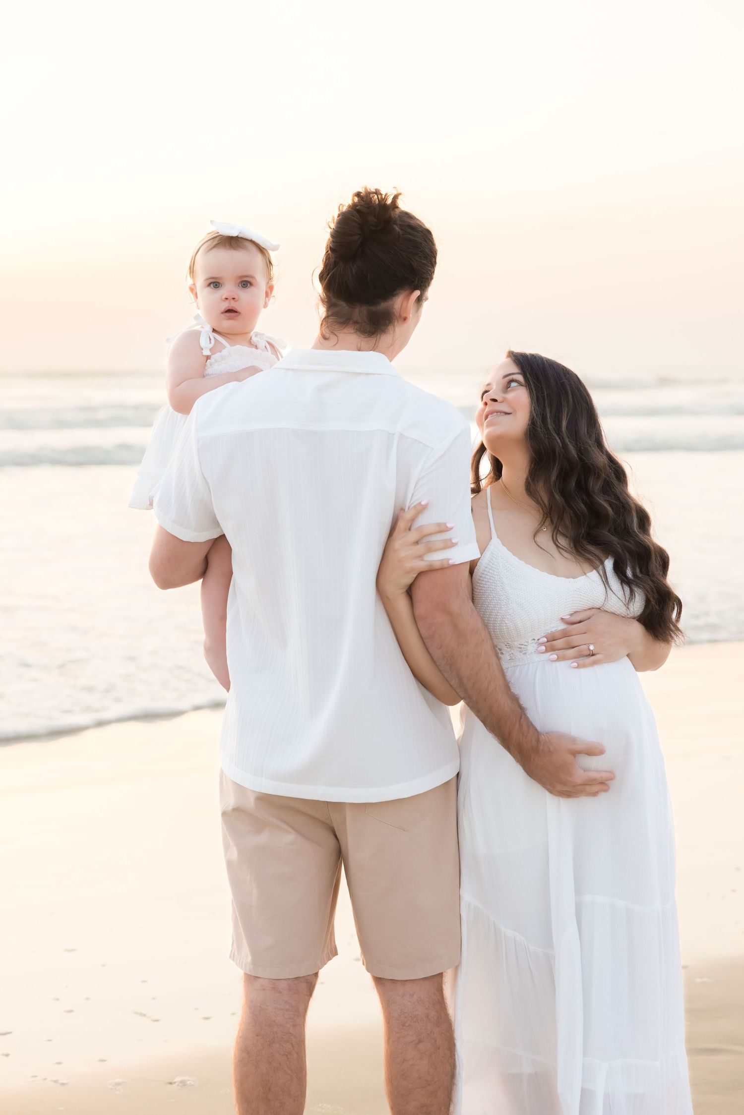 Pregnant woman in white dress with husband holding baby looking toward ocean at sunset.