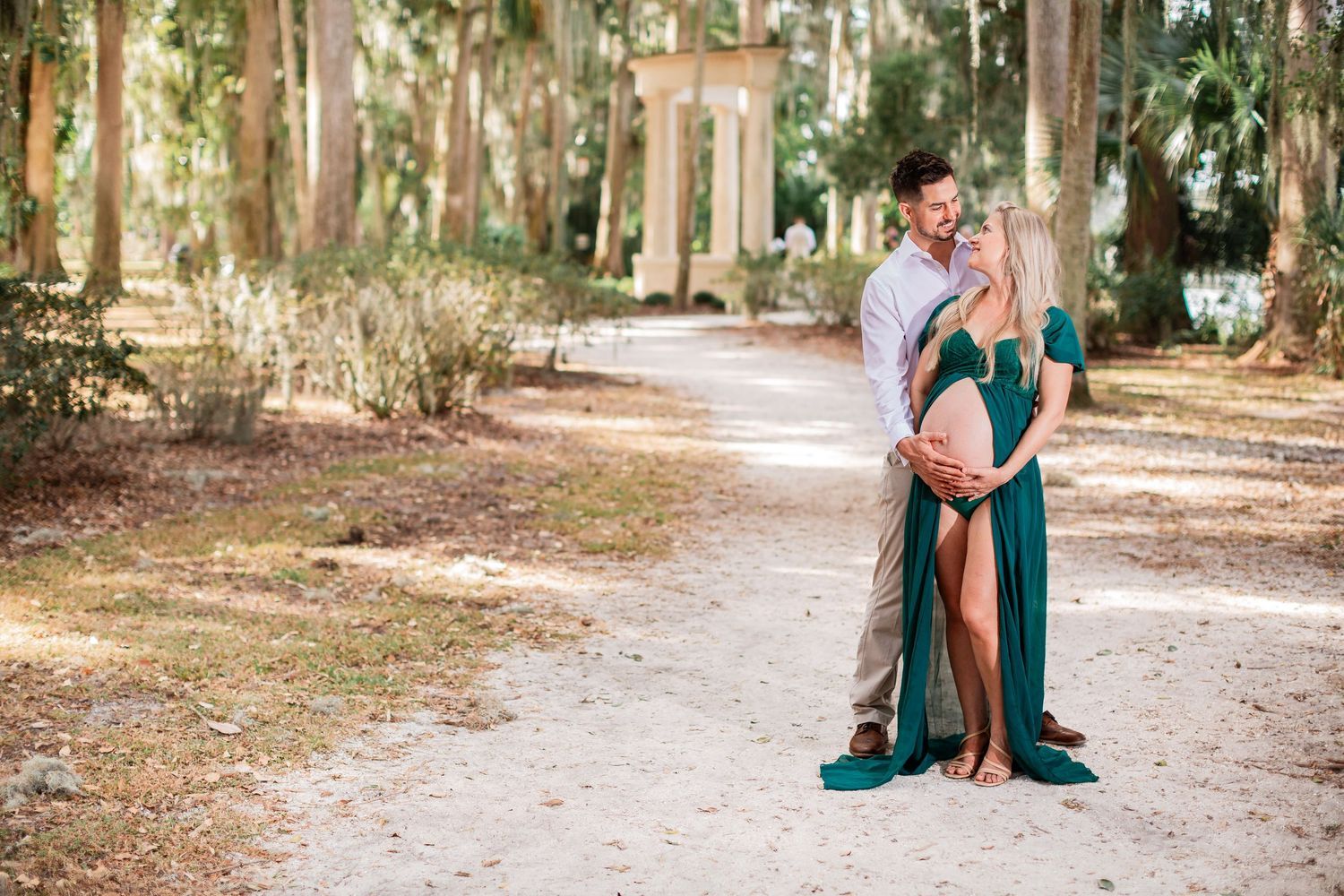 Couple sharing a tender moment on a sandy path surrounded by trees during a maternity photoshoot.
