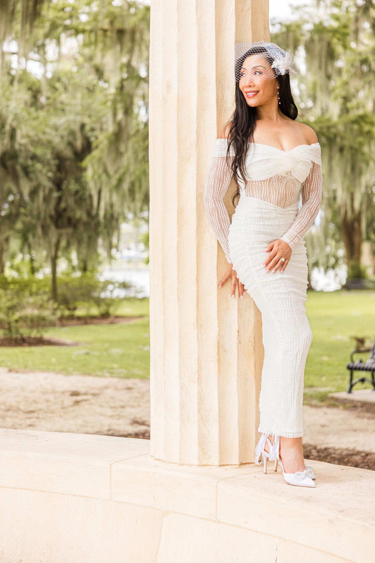Bride in fitted white lace gown and birdcage veil poses by column in lush park.