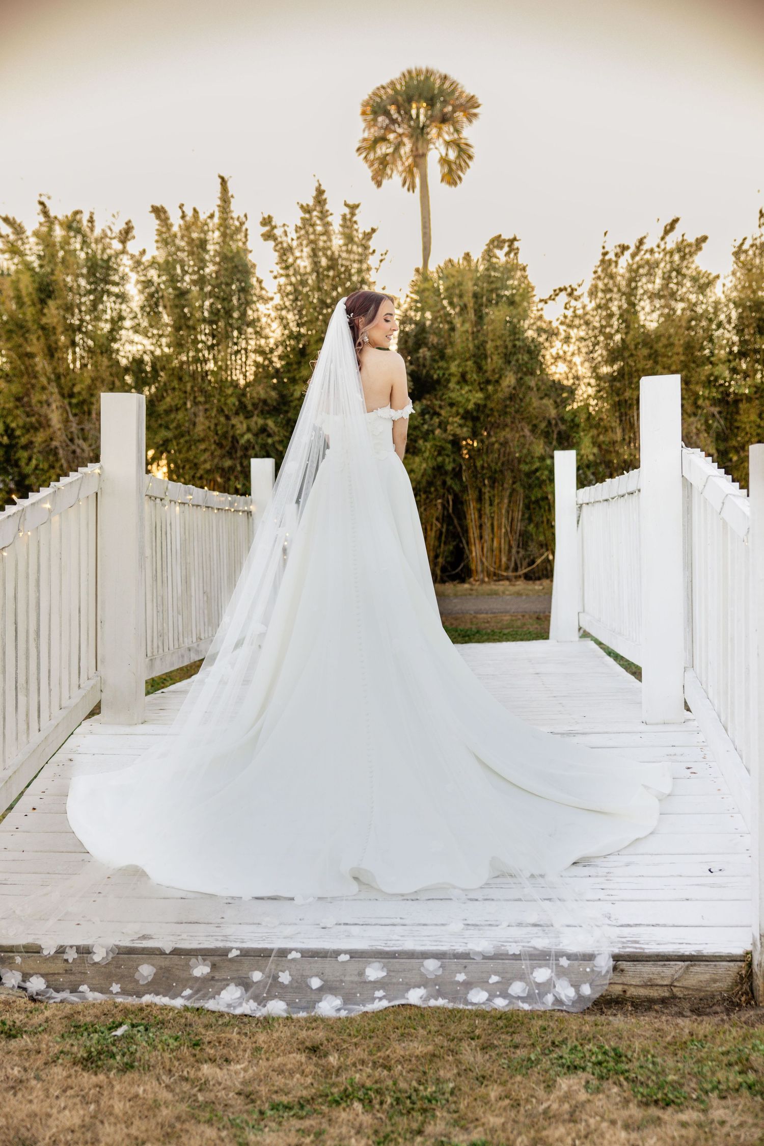 Bride in strapless gown with long floral-trimmed cathedral veil stands on white wooden bridge at sunset.
