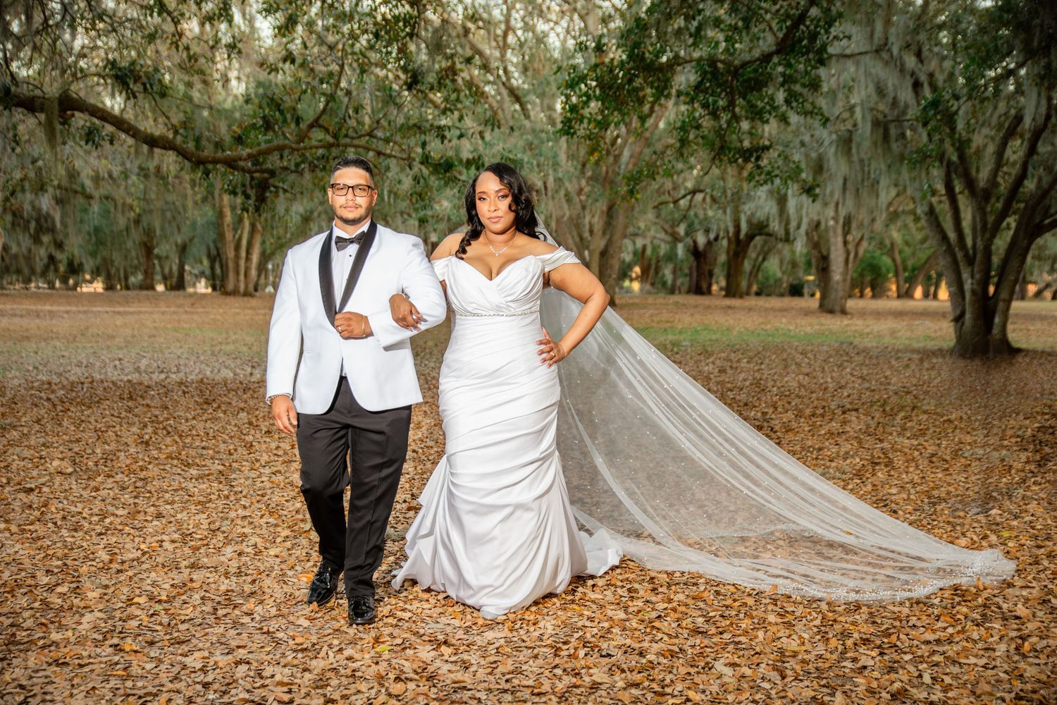 Elegant wedding couple posing under sprawling oak trees on autumn leaf-covered ground at outdoor venue.