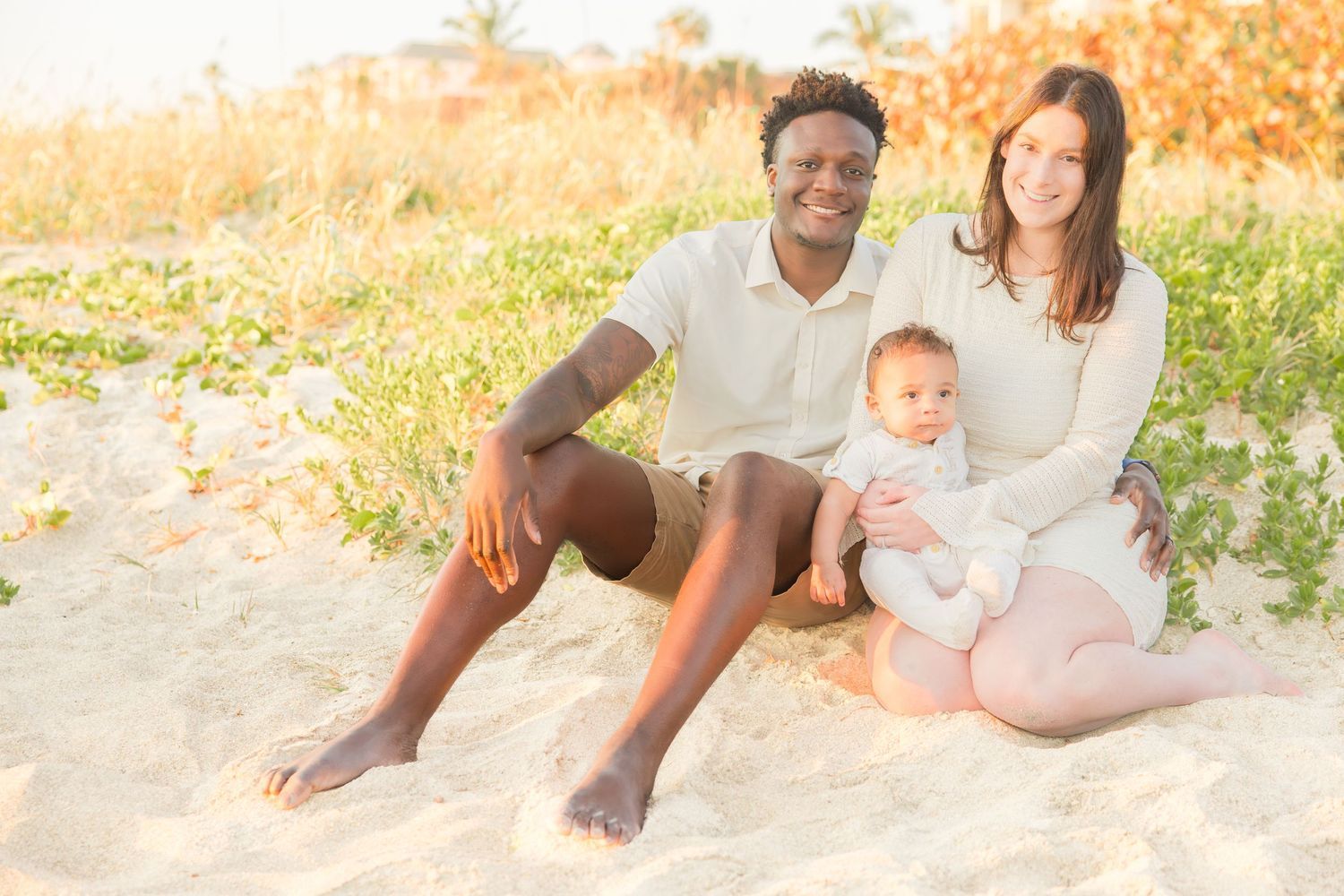 Young interracial family with baby sitting together on sandy beach at golden hour.