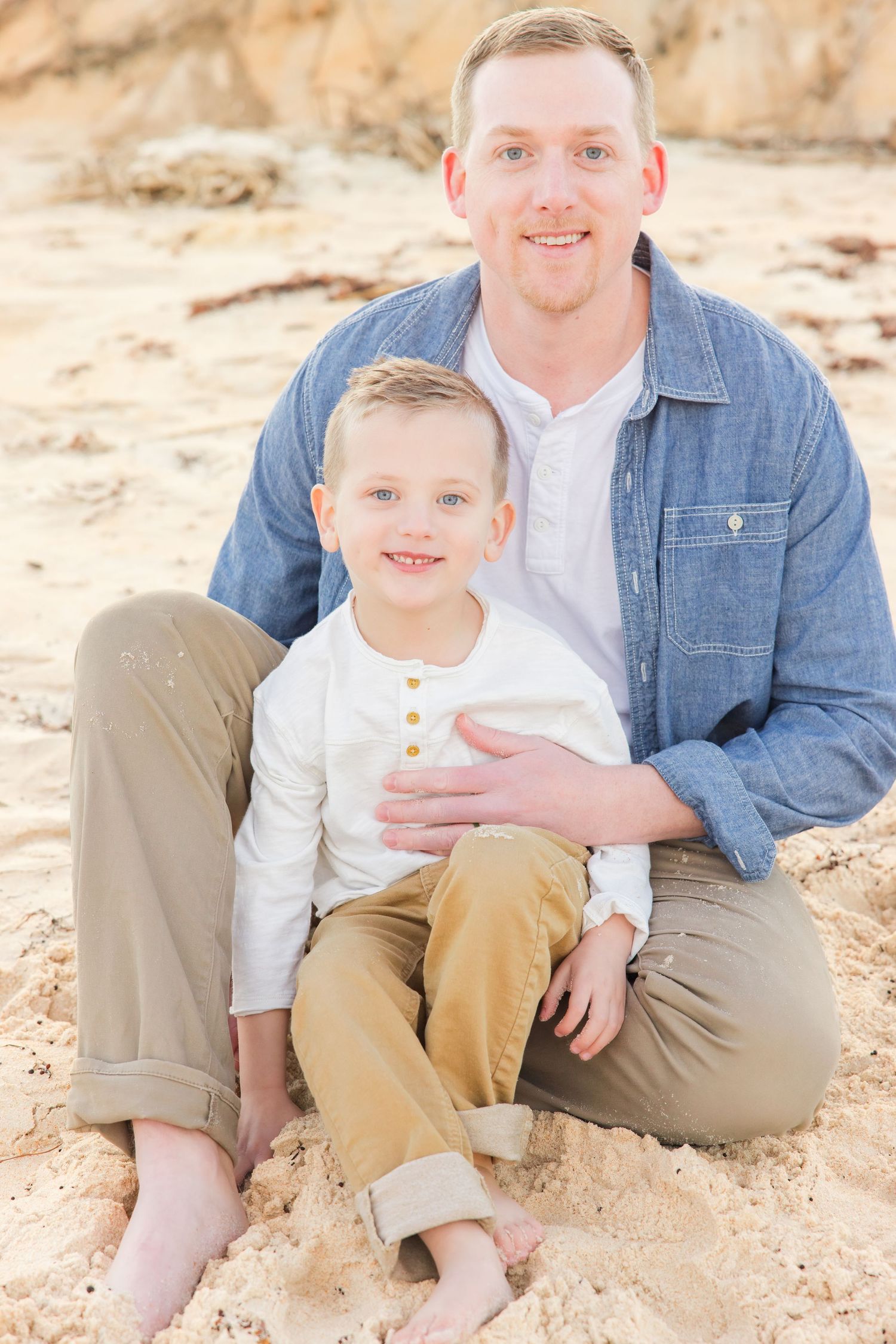 Smiling father sitting on sandy beach with his young blond son in a warm outdoor portrait.