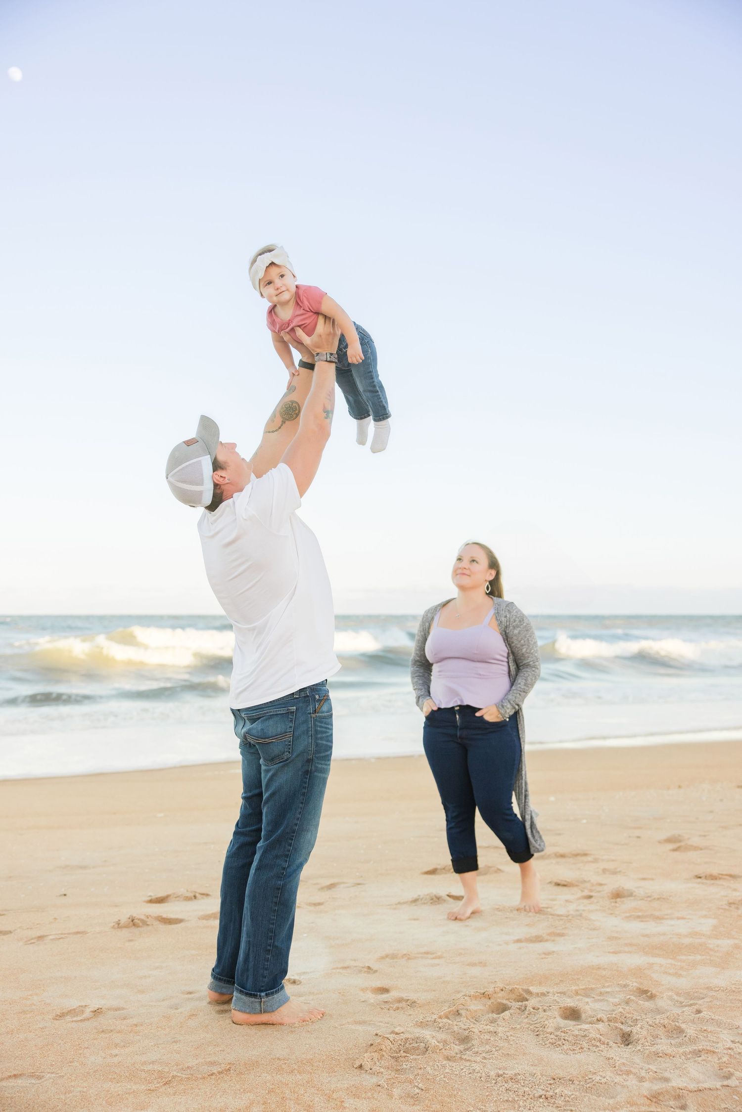 Father tossing toddler in air on beach while mother watches smiling.
