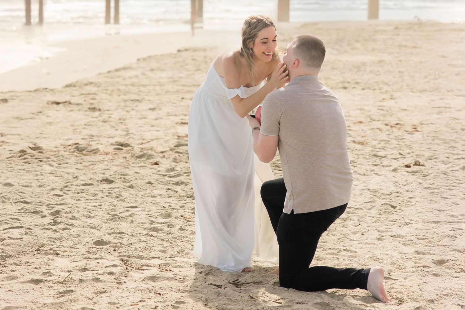 Man proposing on one knee to woman in white dress on sandy beach at sunset.