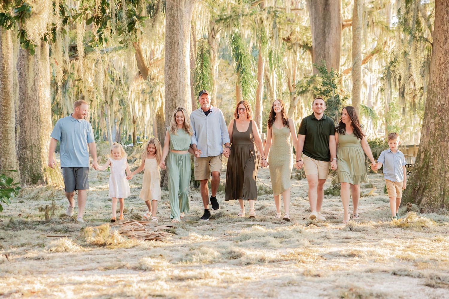 Extended family of ten walking hand-in-hand through a sunlit moss-draped forest path.