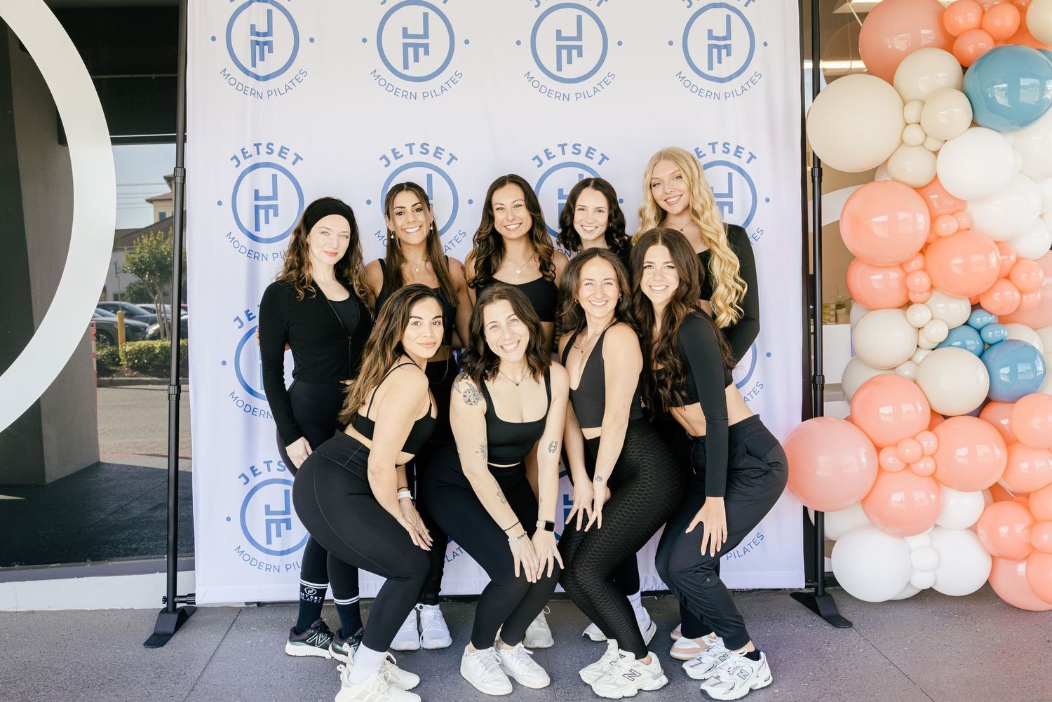 Group of nine women in black athletic wear pose at fitness studio grand opening event.