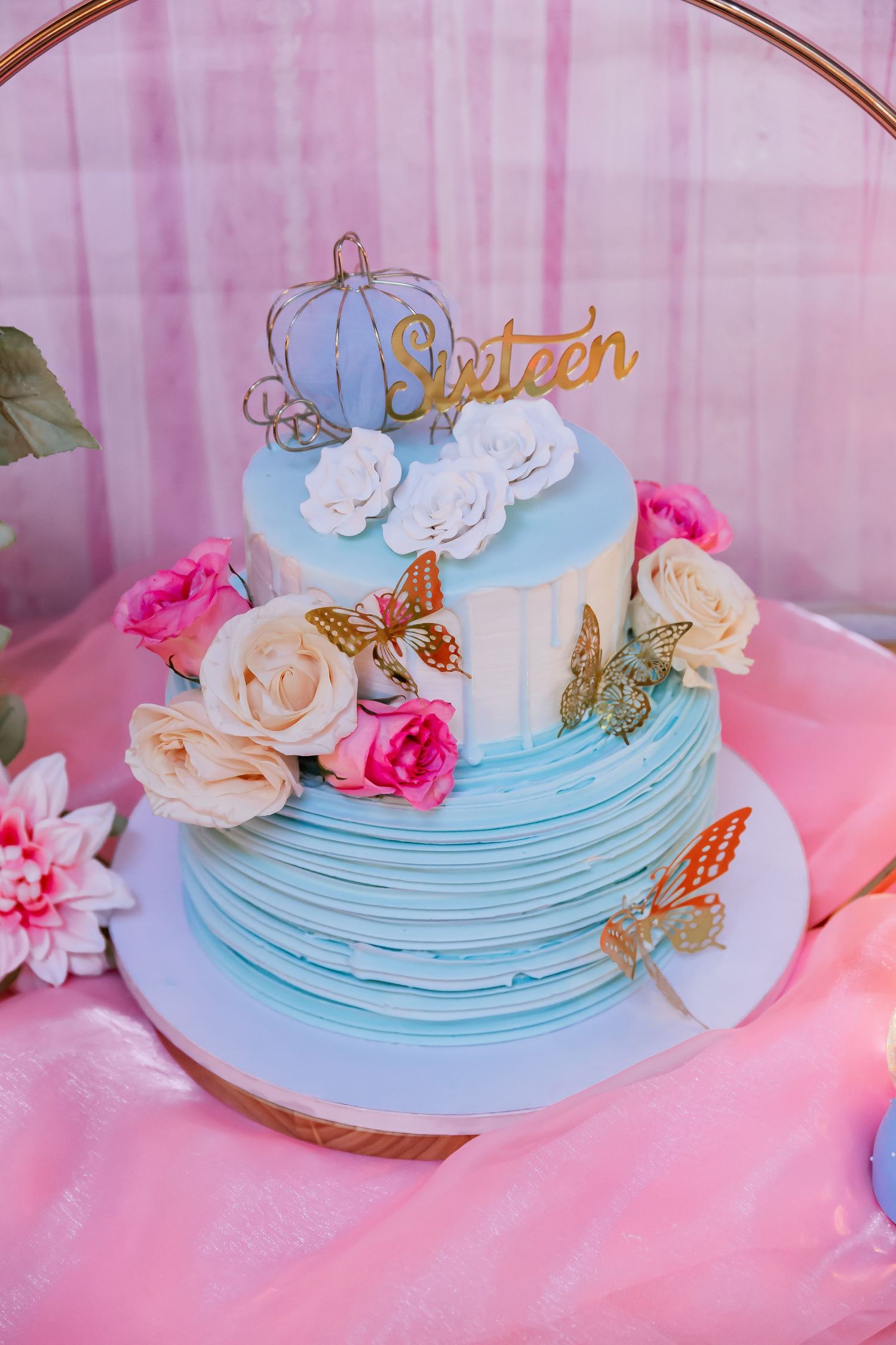 Two-tier light blue Sweet Sixteen birthday cake with roses, butterflies, and carriage topper.