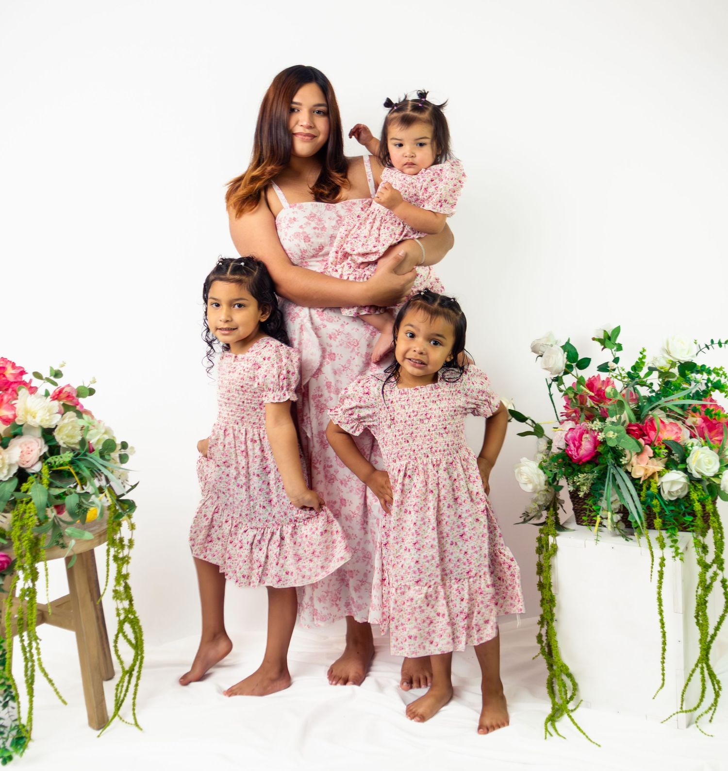 A mother and three daughters pose in matching pink floral dresses with flower arrangements nearby.