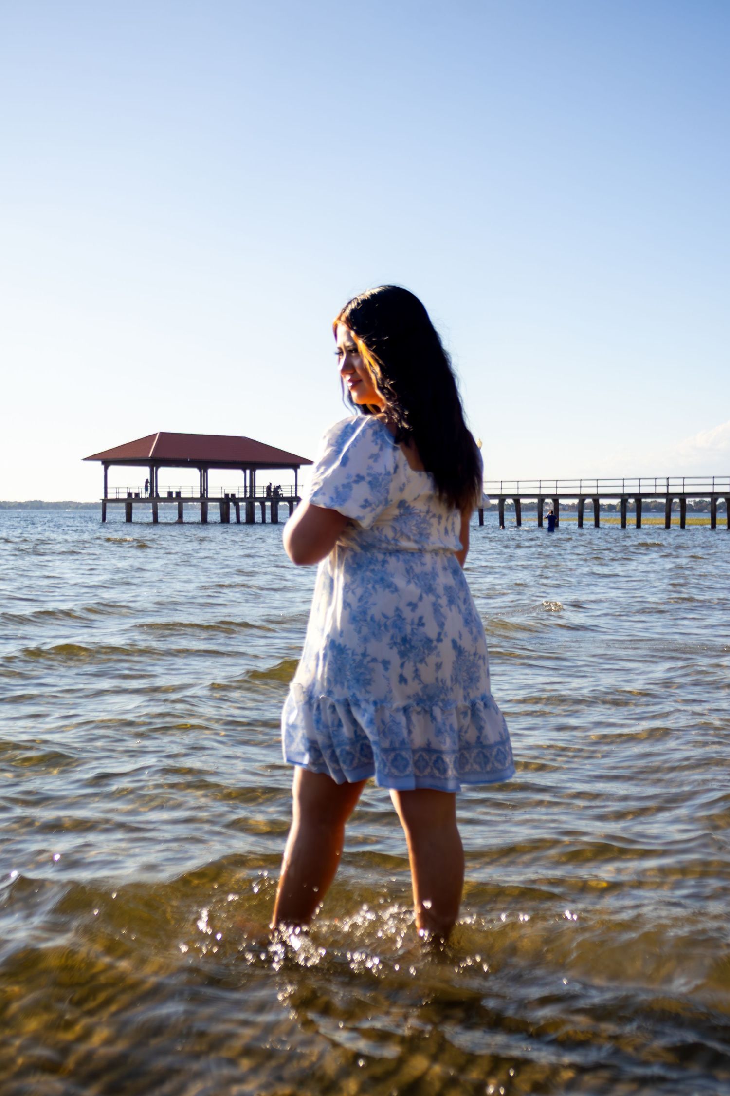Young woman in blue floral dress standing in ocean water near pier at golden hour sunset.