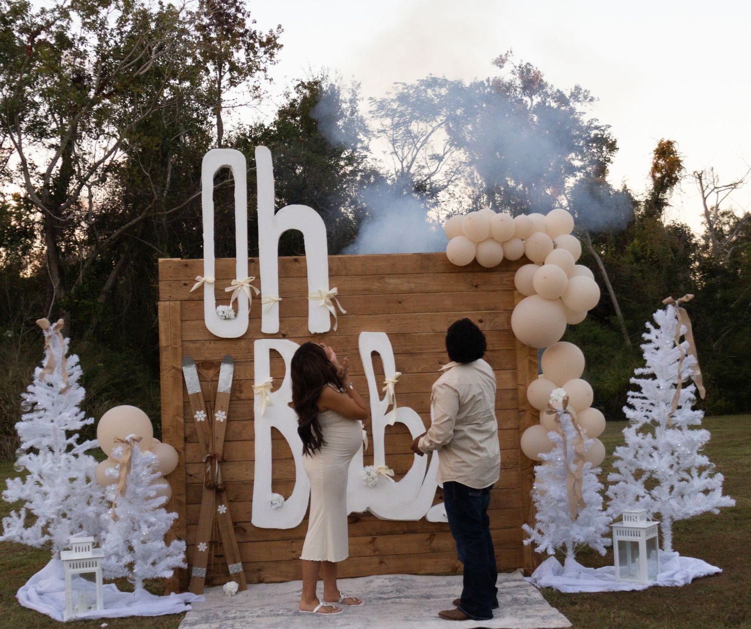 A couple reveals baby gender with blue smoke at an outdoor Oh Baby decorated wooden backdrop.