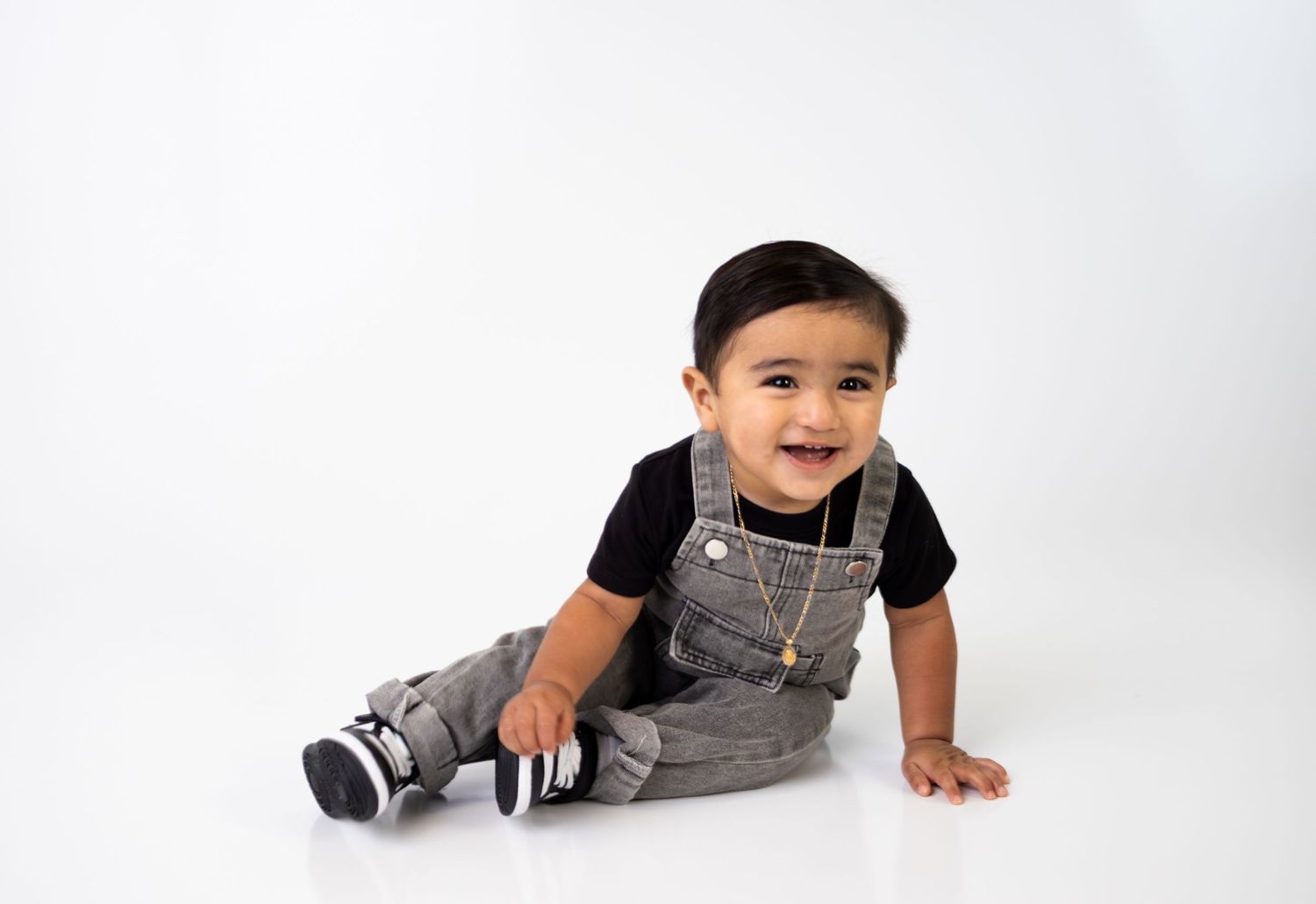 Happy toddler in gray overalls and black shirt with gold necklace on white background.