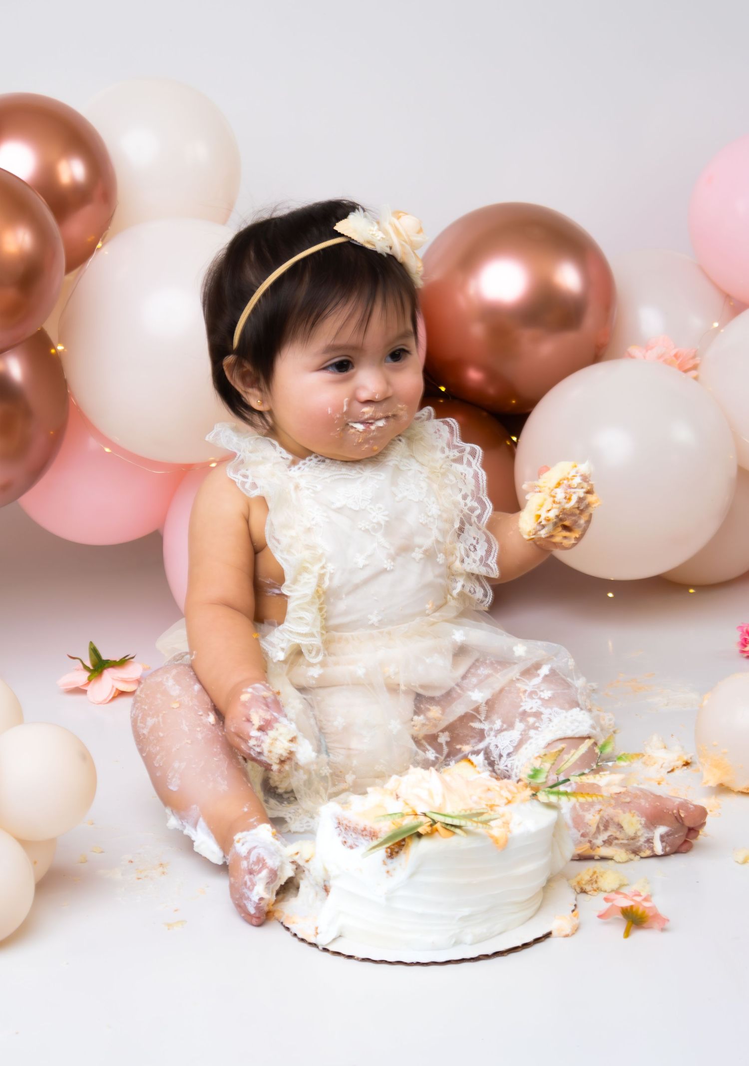 Baby girl in lace dress smashes first birthday cake surrounded by pink and gold balloons.