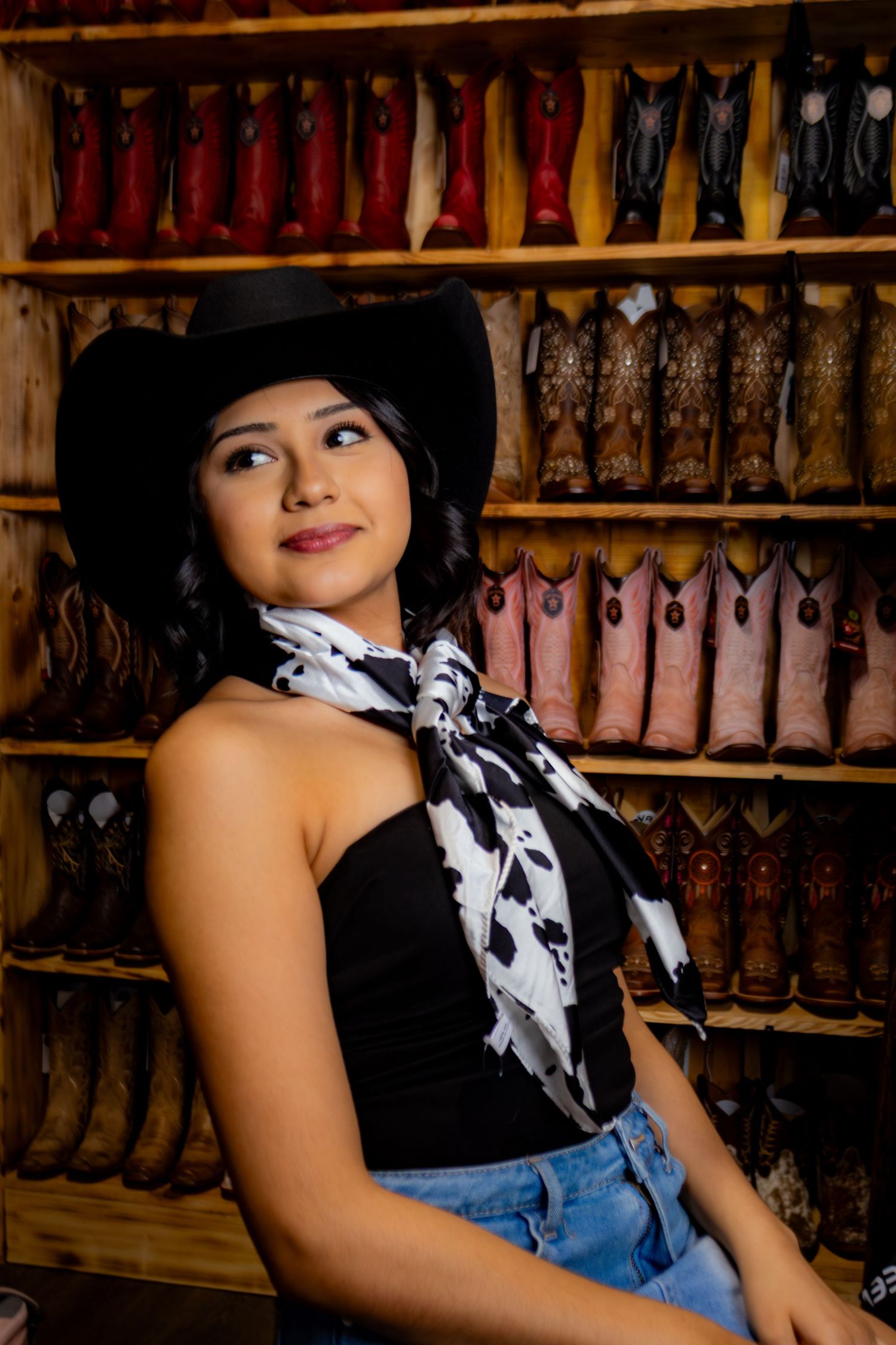 Young woman in black cowboy hat and cow print scarf posing in a western boot store.