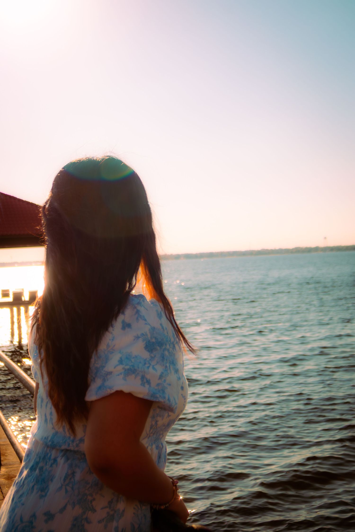 Young woman in blue floral dress gazing at ocean from pier during bright sunny day.