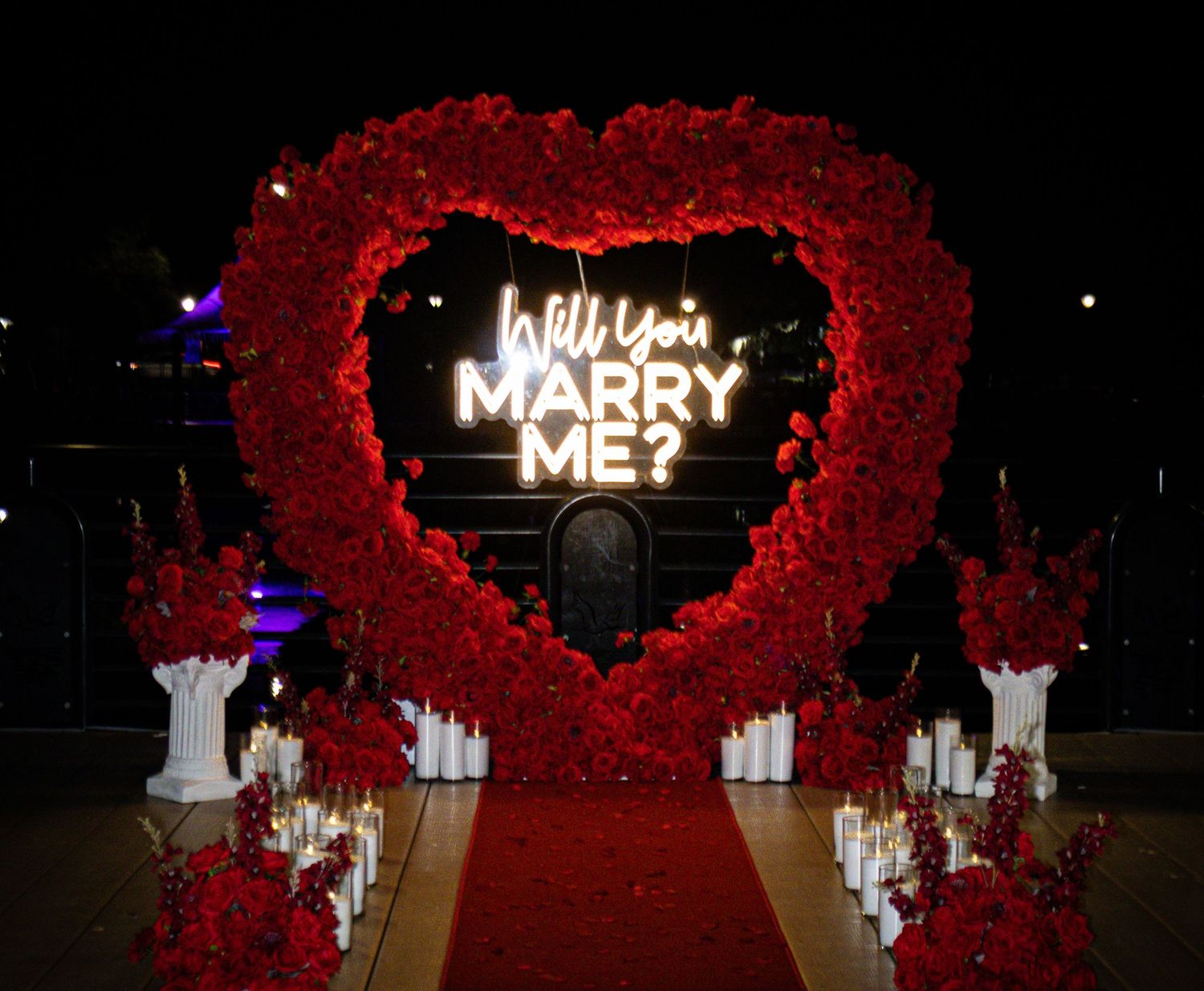 Romantic red rose heart arch with Will You Marry Me neon sign and candles at night.