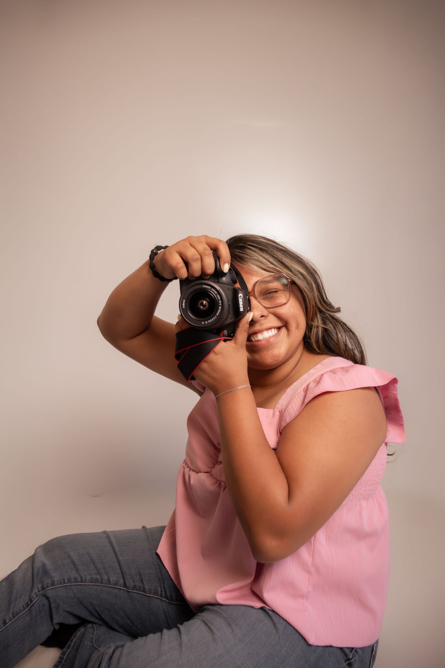 Smiling woman in pink top holding a Canon DSLR camera up to her eye, sitting against a light background.