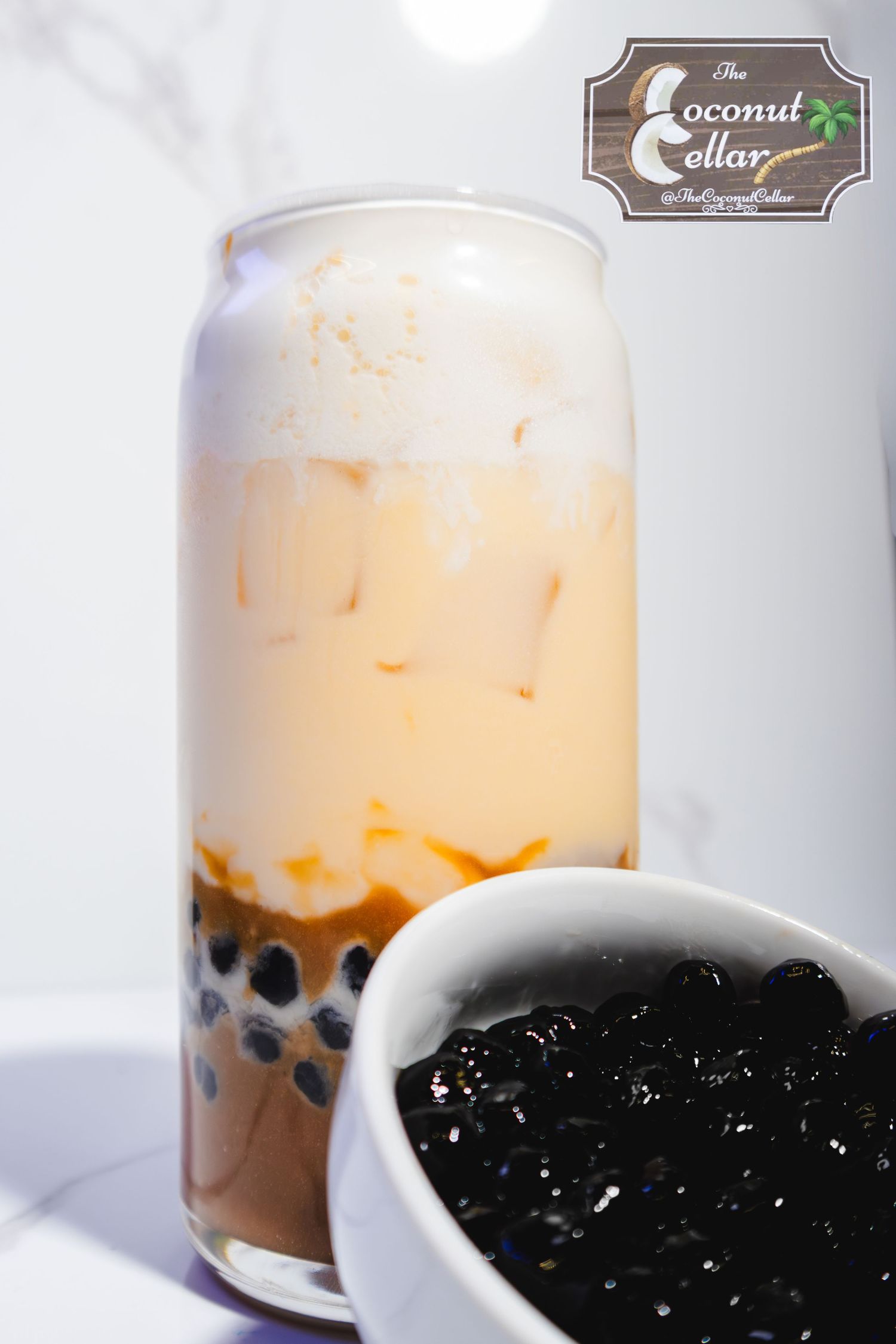 The Coconut Cellar creamy layered boba tea with black tapioca pearls in tall glass can on white surface.