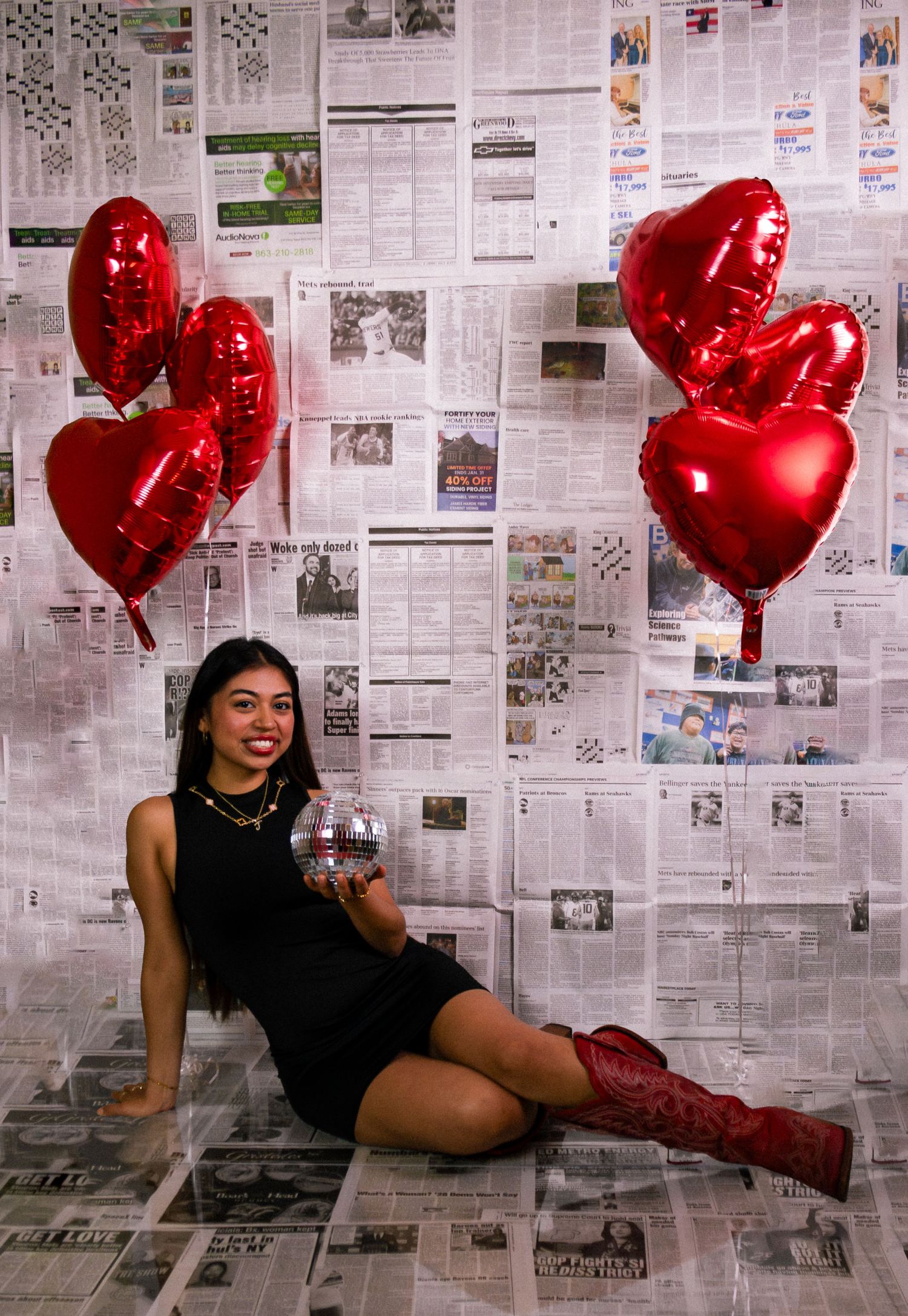 Young woman in black dress and red cowboy boots poses with disco ball and heart balloons on newspaper backdrop.