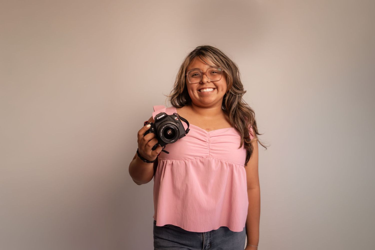 Smiling young woman in pink top holding a DSLR camera, posing against a neutral gray background.