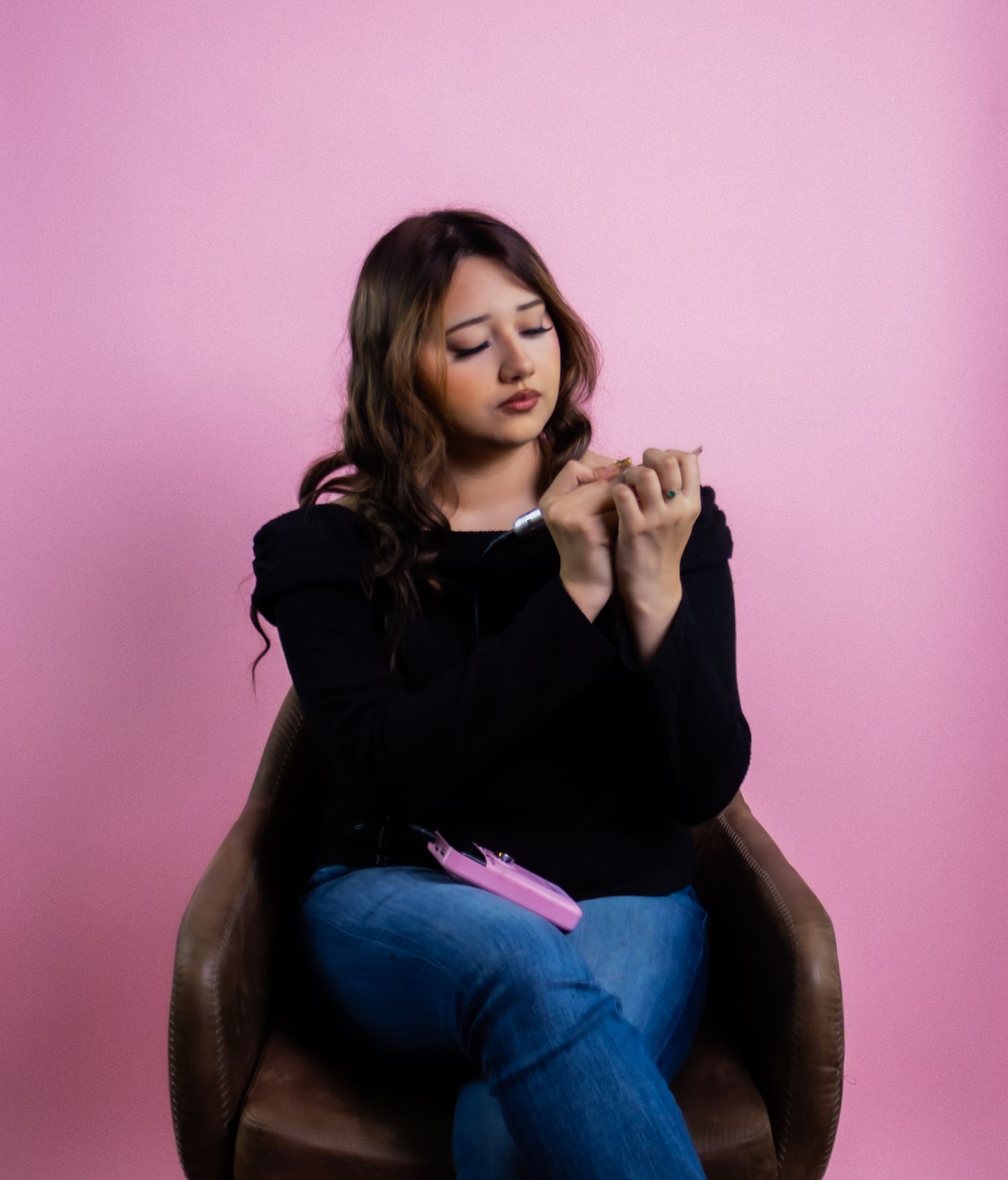 Young woman in black sweater and jeans sitting in brown chair examining her nails on pink background.