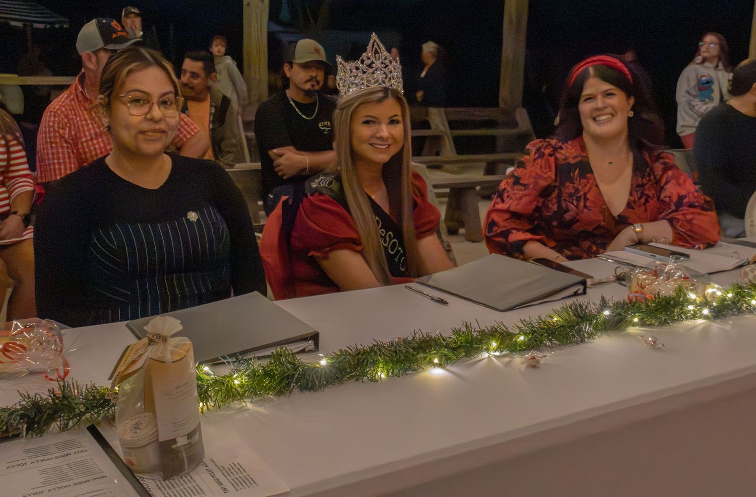 Three women smile while seated at a judging table decorated with garland lights at an indoor pageant event.