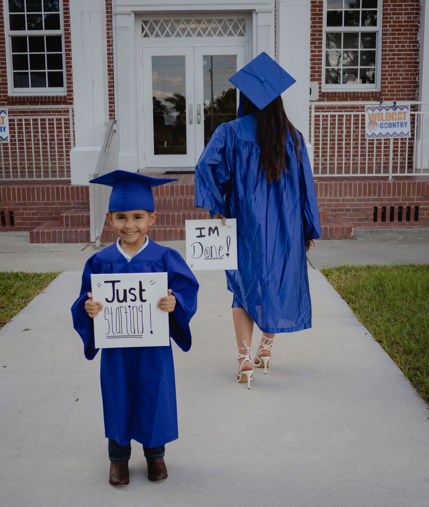 Two children in blue graduation caps and gowns hold signs outside a brick building, celebrating a milestone together.