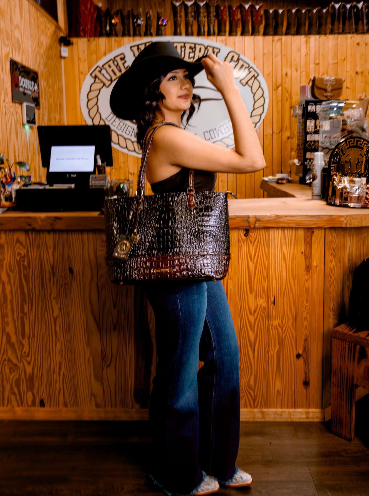 Woman modeling black crocodile tote bag and black cowboy hat in The Western Cut store.
